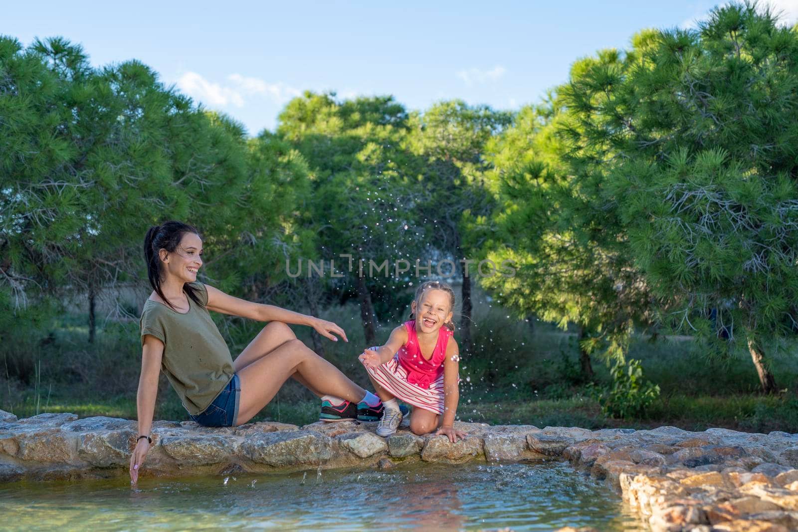 Beautiful mother and her little daughter outdoors. Beauty Mum and her Child playing with water in Park together at sunset. Outdoor Portrait of happy family. Mother's Day.