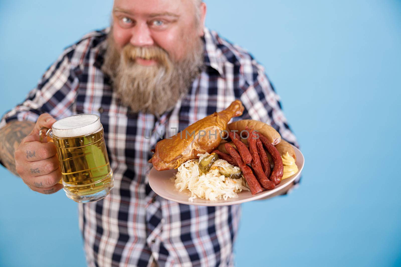 Smiling obese man stands on light blue background in studio, focus on hands with plate of greasy food and mug of beer