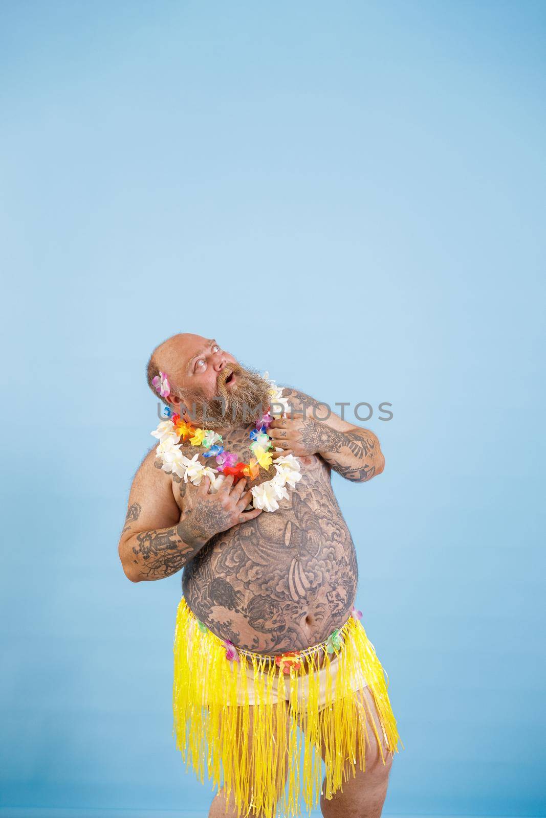 Surprised middle aged plump man in decorative yellow grass skirt and flowers garland looks up posing on light blue background in studio