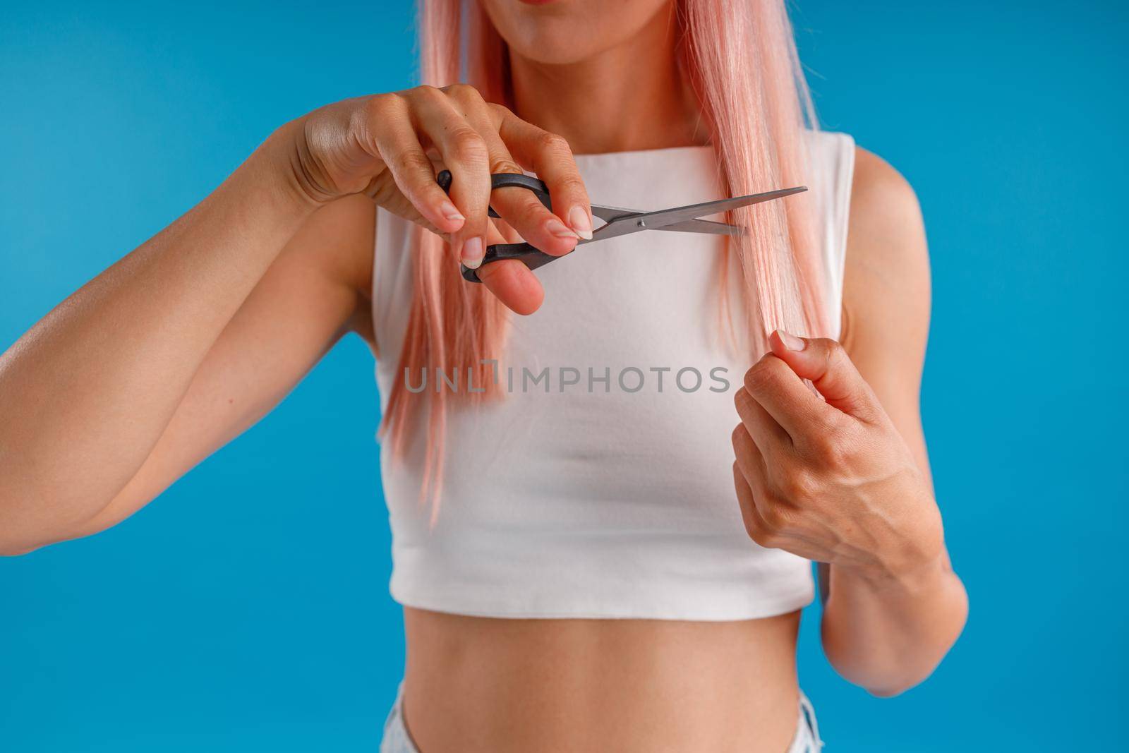 Cropped shot of woman with pink hair cutting her hair using scissors, standing isolated over blue studio background. Beauty and hair care concept