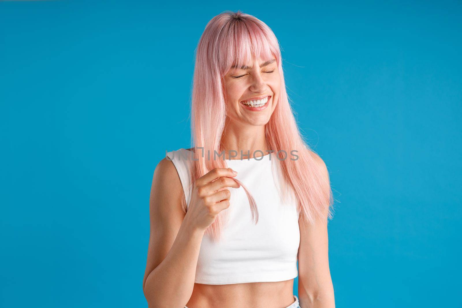 Joyful young woman with natural long pink dyed hair laughing with eyes closed, posing isolated over blue studio background. Beauty, hair care concept