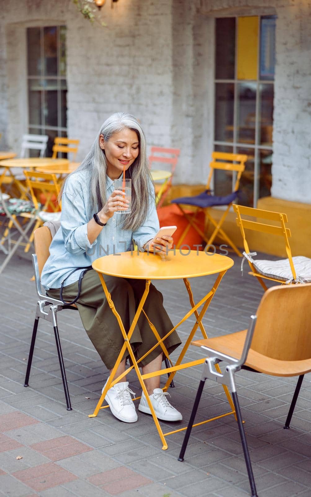 Positive senior Asian lady drinks water and uses mobile phone on outdoors cafe terrace by Yaroslav_astakhov