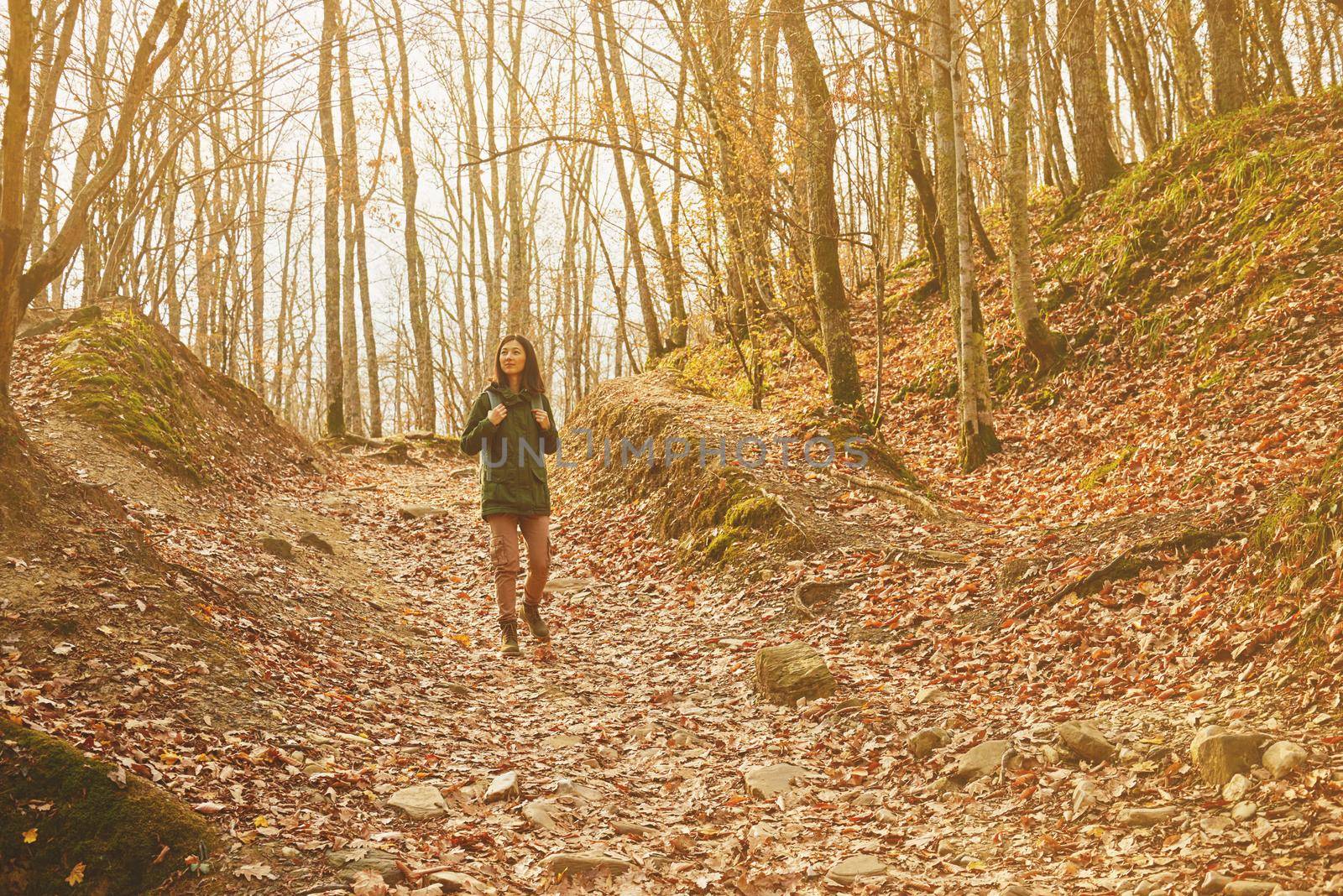 Smiling hiker woman walking in beautiful autumn forest, dry yellow leaves on land. Hiking and leisure theme. Image with sunlight effect