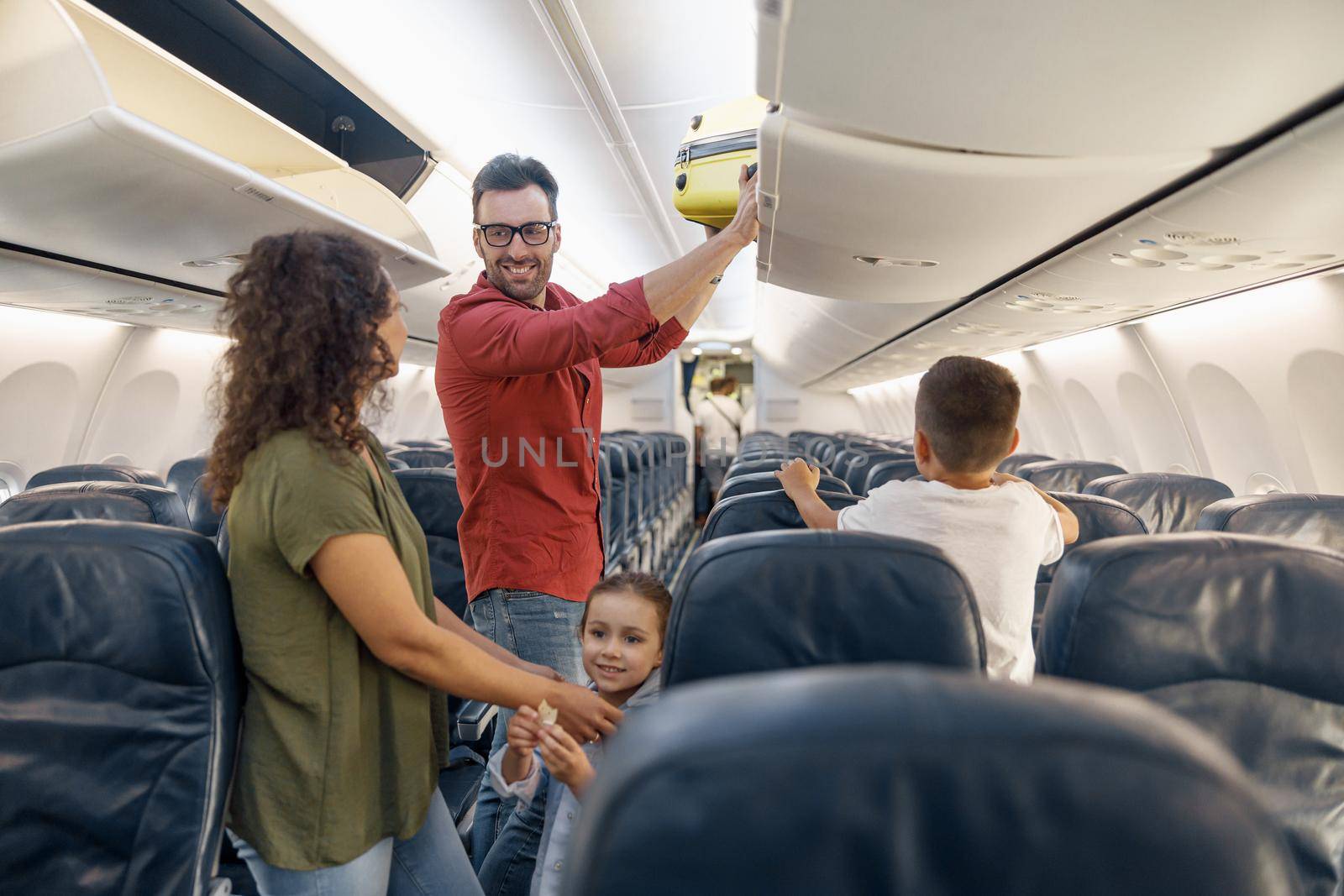 Cheerful man looking at his wife with a smile and putting carry on luggage in compartment while traveling together with his family by plane by Yaroslav_astakhov
