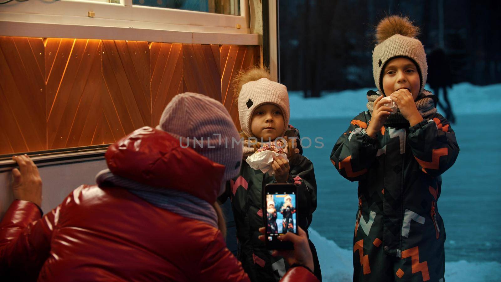 A young woman taking photos of her children drinking hot drinks and eating donuts outdoors in winter. Mid shot