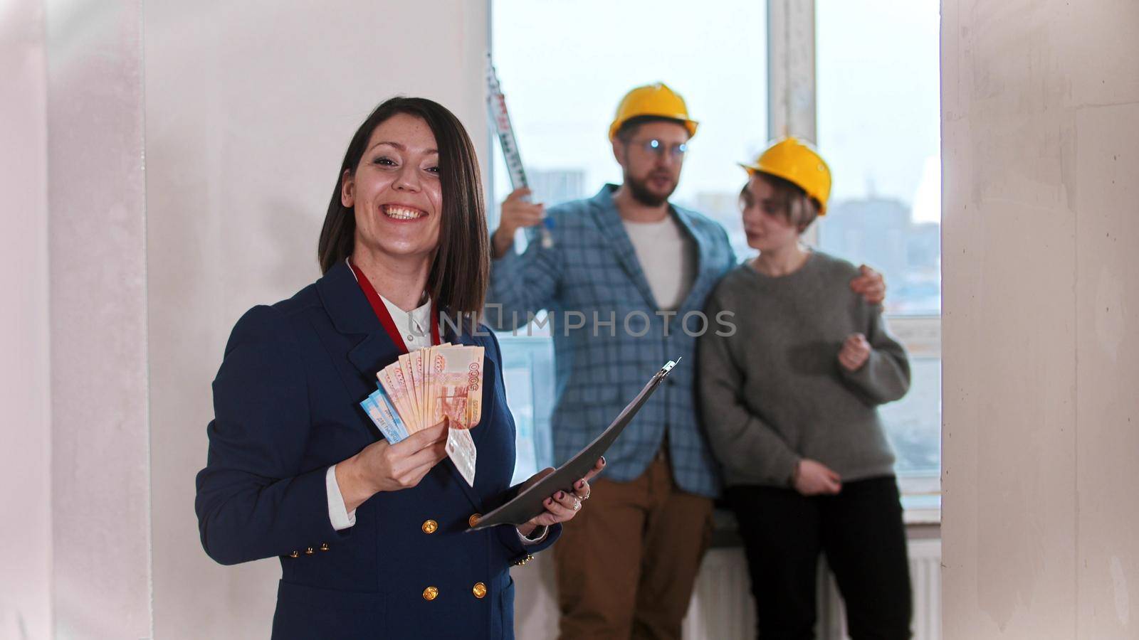 A real estate agent holding a lot of money in cash - young married couple looking around on the background. Mid shot