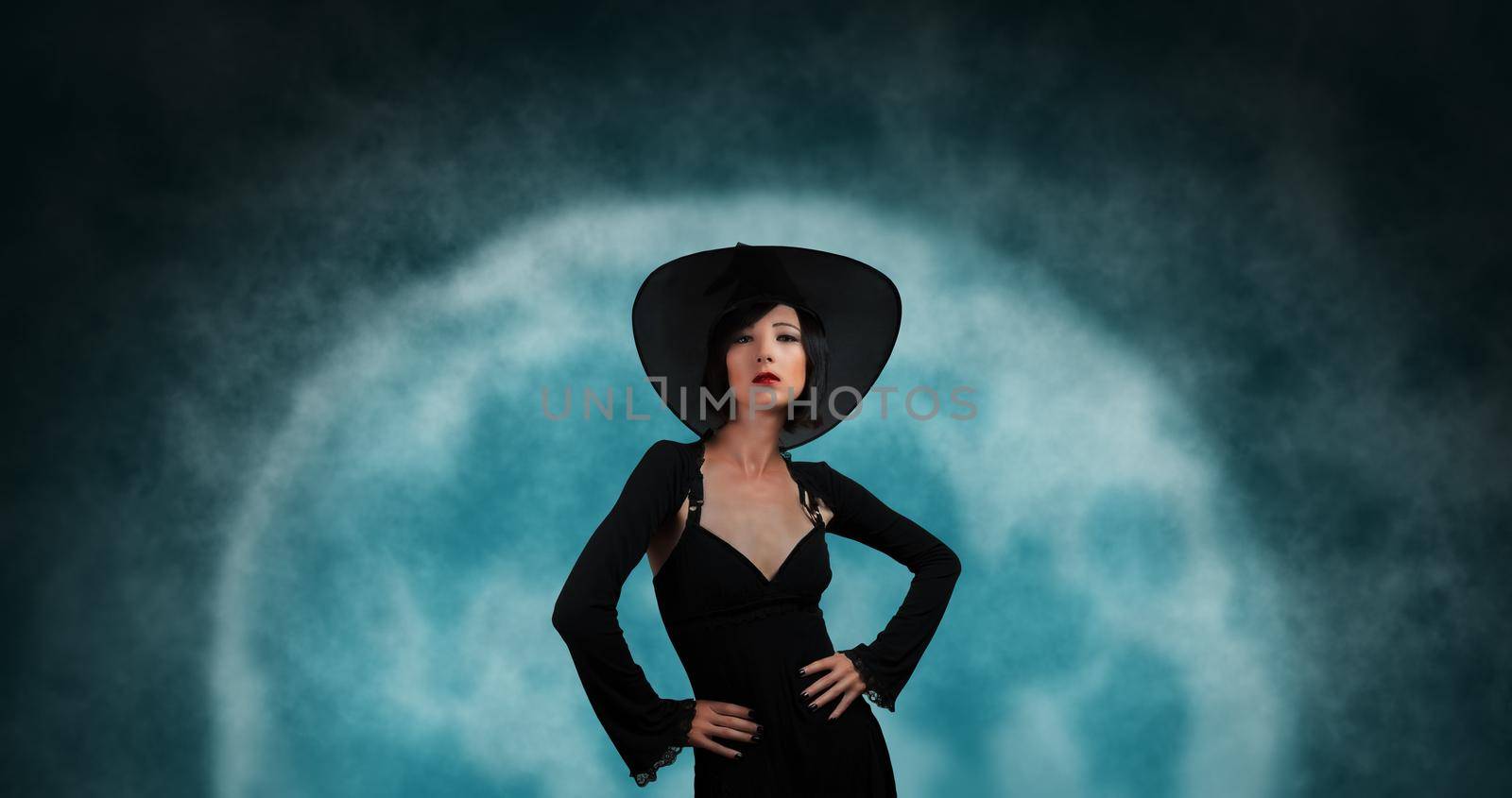 Attractive fashionable witch in black hat and dress on background of full moon. Halloween theme