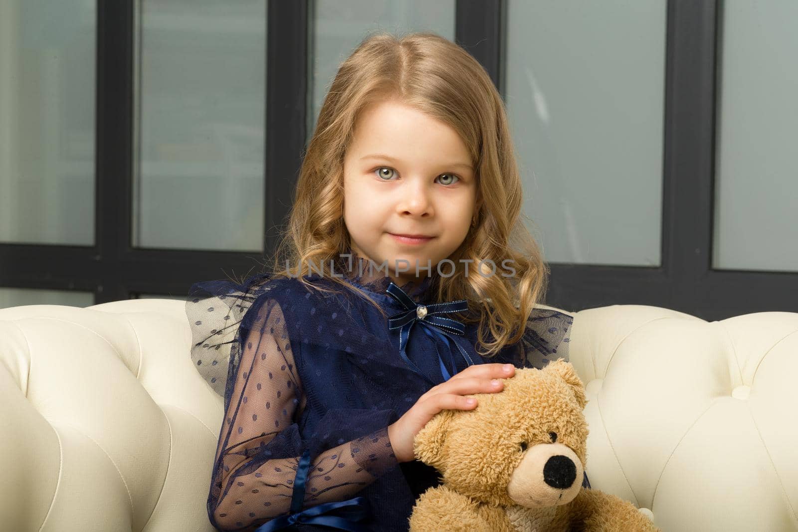 Lovely little girl hugging her teddy bear toy. Adorable blonde girl wearing trendy clothes sitting on white leather comfortable sofa and smiling at camera.