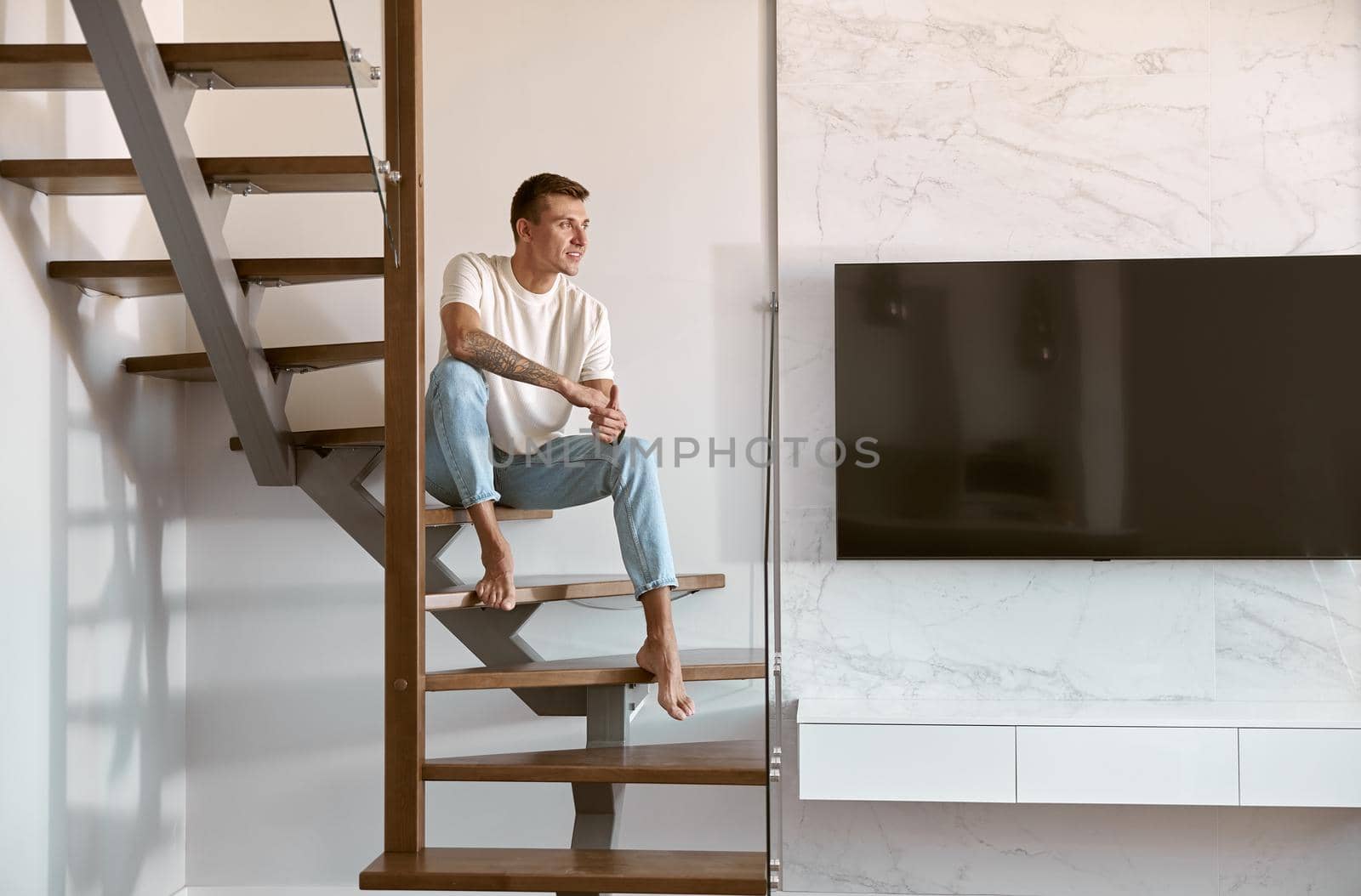 Relaxing happy caucasian man on the minimalistic stairs in modern light apartment by Yaroslav_astakhov