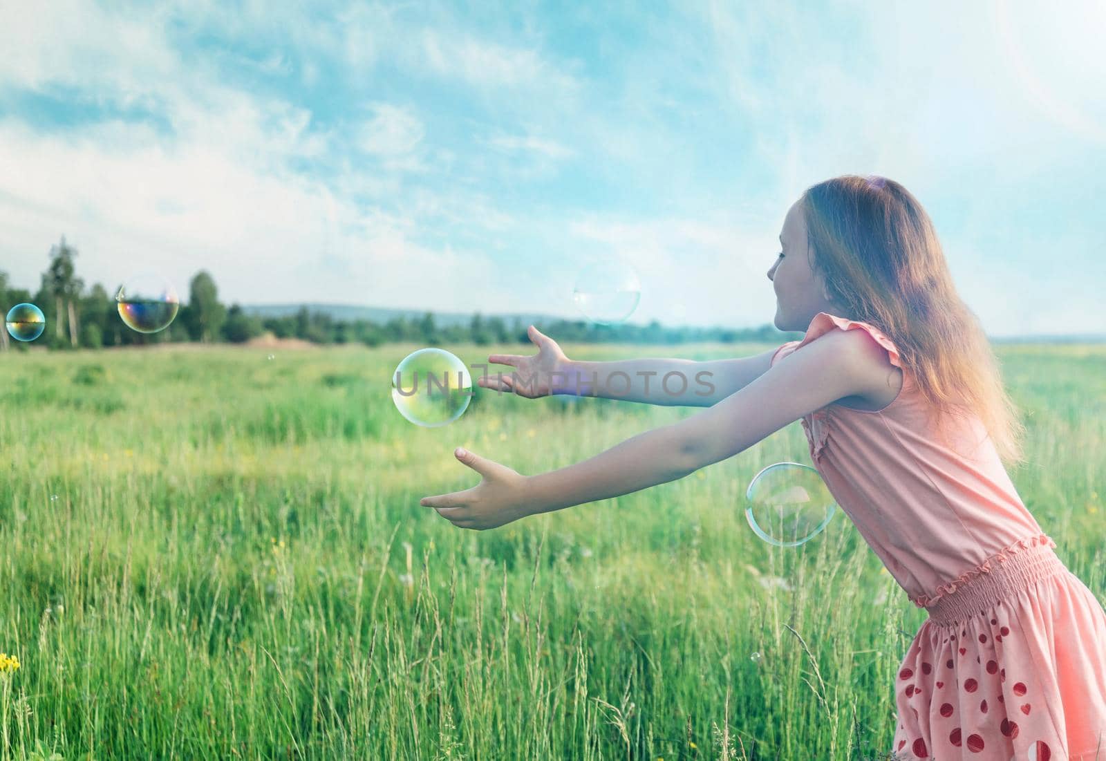 Cheerful little girl playing among soap bubbles on summer meadow outdoor. Concept of happiness and carefree