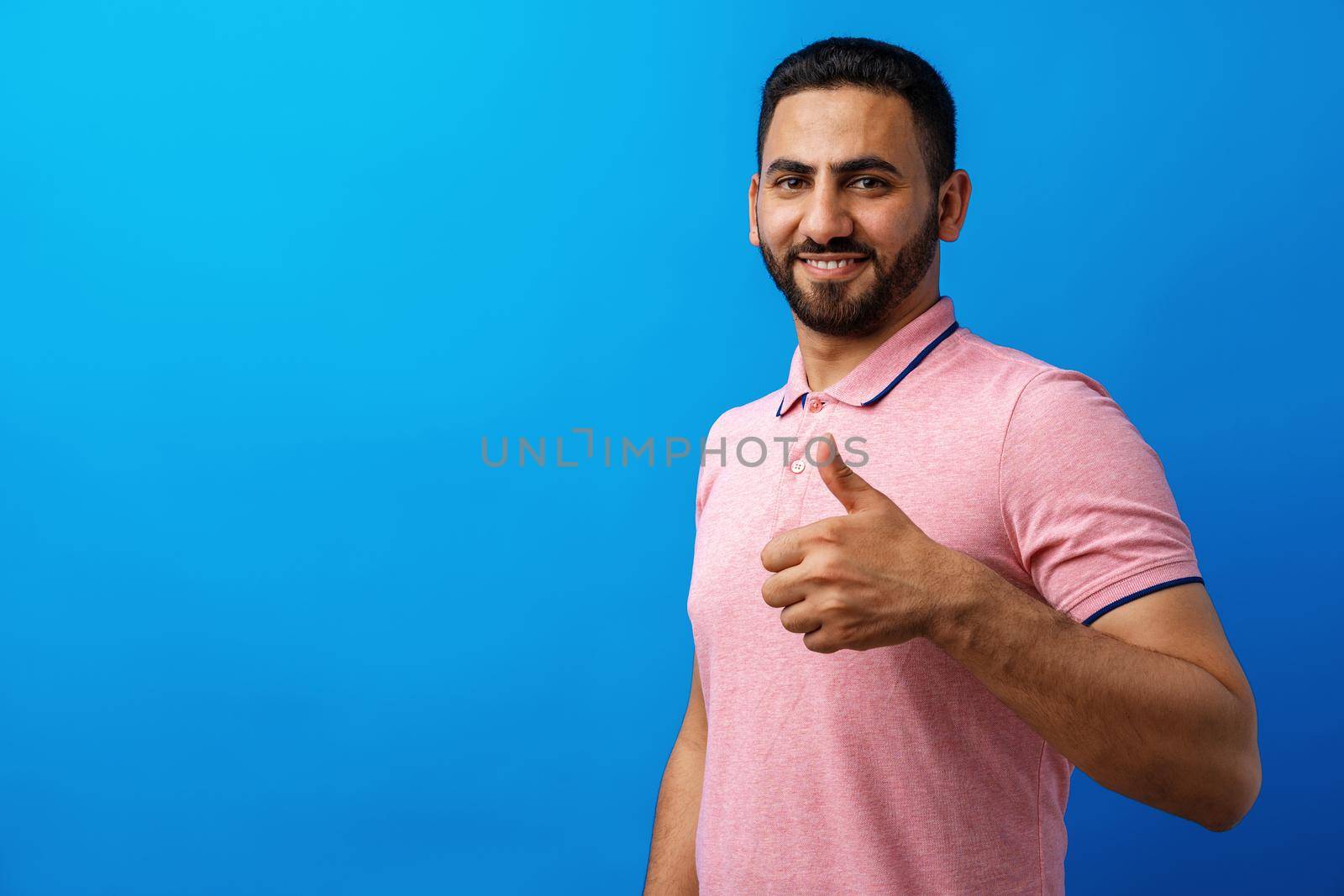 Happy beard young man smiling with thumbs up against blue background, close up