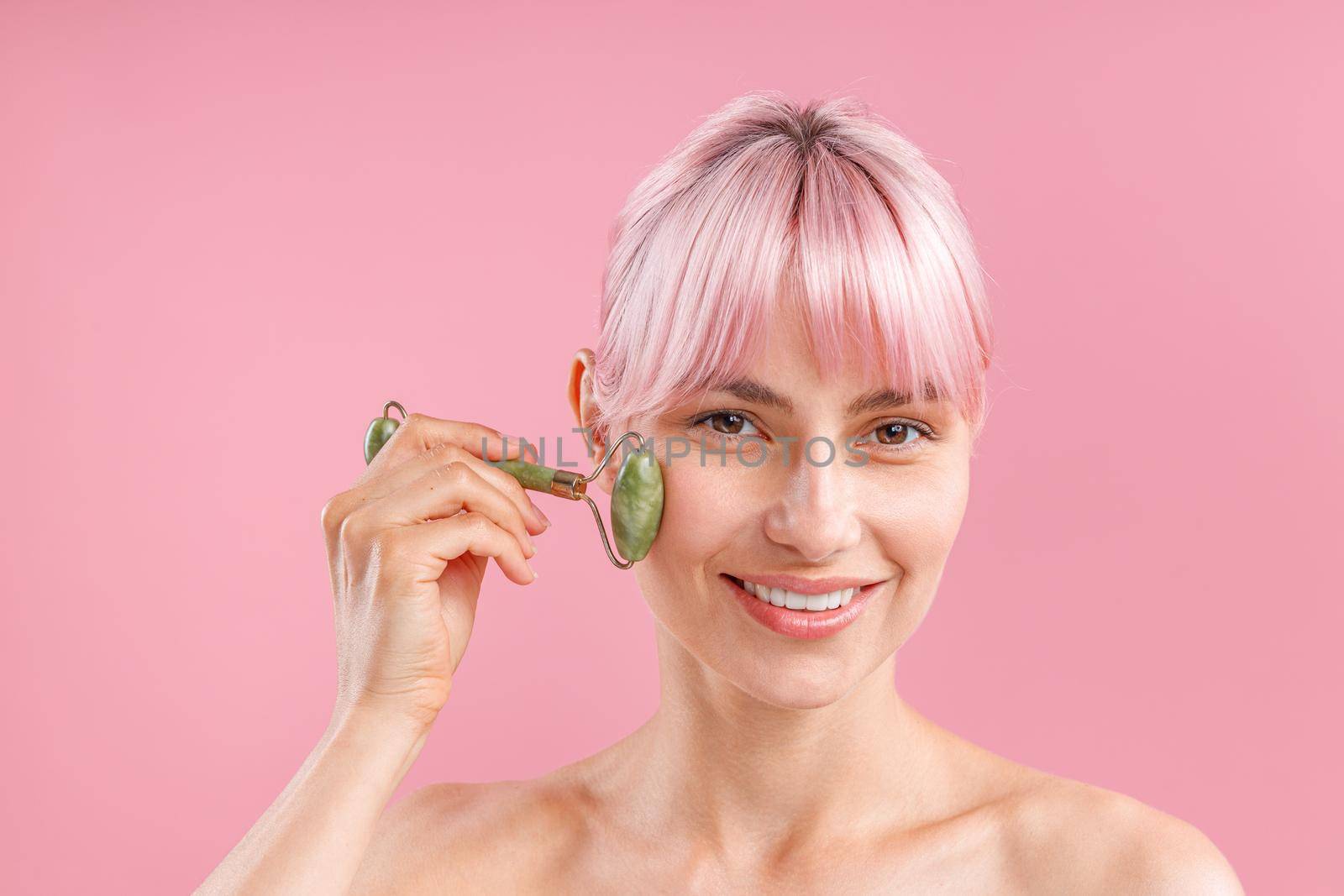 Cheerful naked woman with pink hair smiling at camera while using natural jade facial roller for skin care isolated over pink background by Yaroslav_astakhov