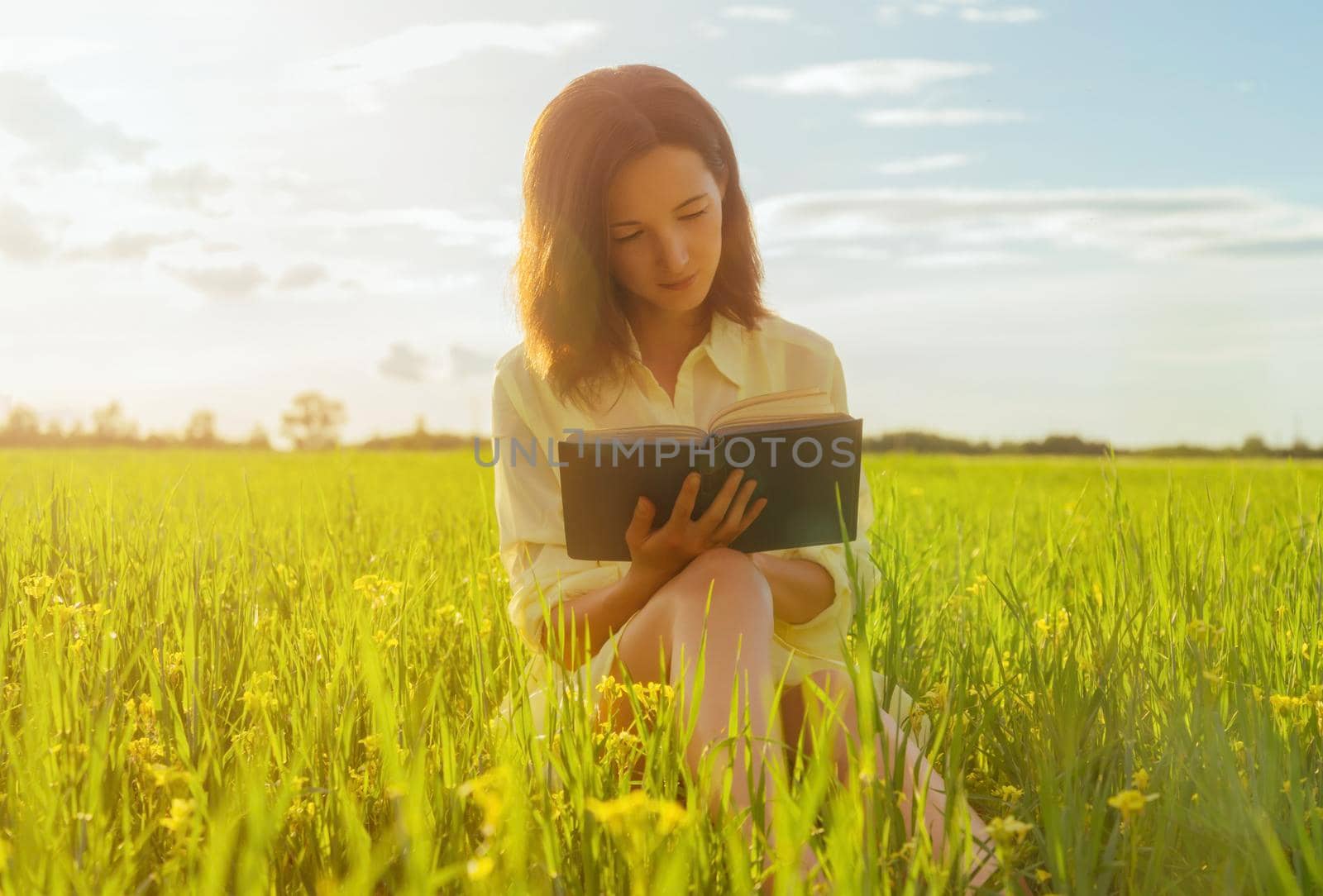 Beautiful young woman reading a book on summer meadow. Image with sunlight effect