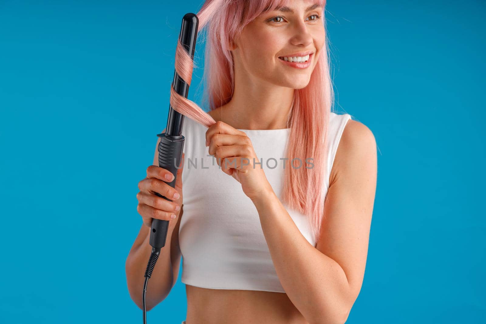 Beautiful smiling woman styling pink long straight hair using curling iron, standing isolated over blue studio background by Yaroslav_astakhov