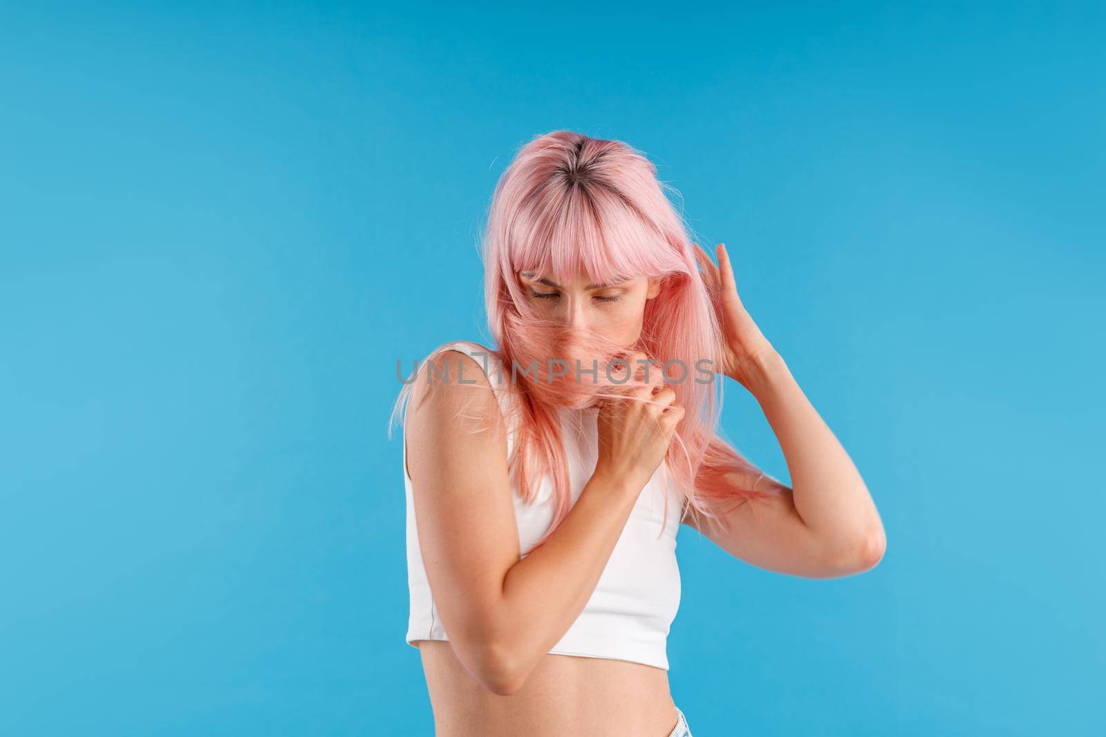 Attractive female model in white shirt looking down, hiding her face with pink hair while posing isolated over blue studio background. Beauty, hair care concept