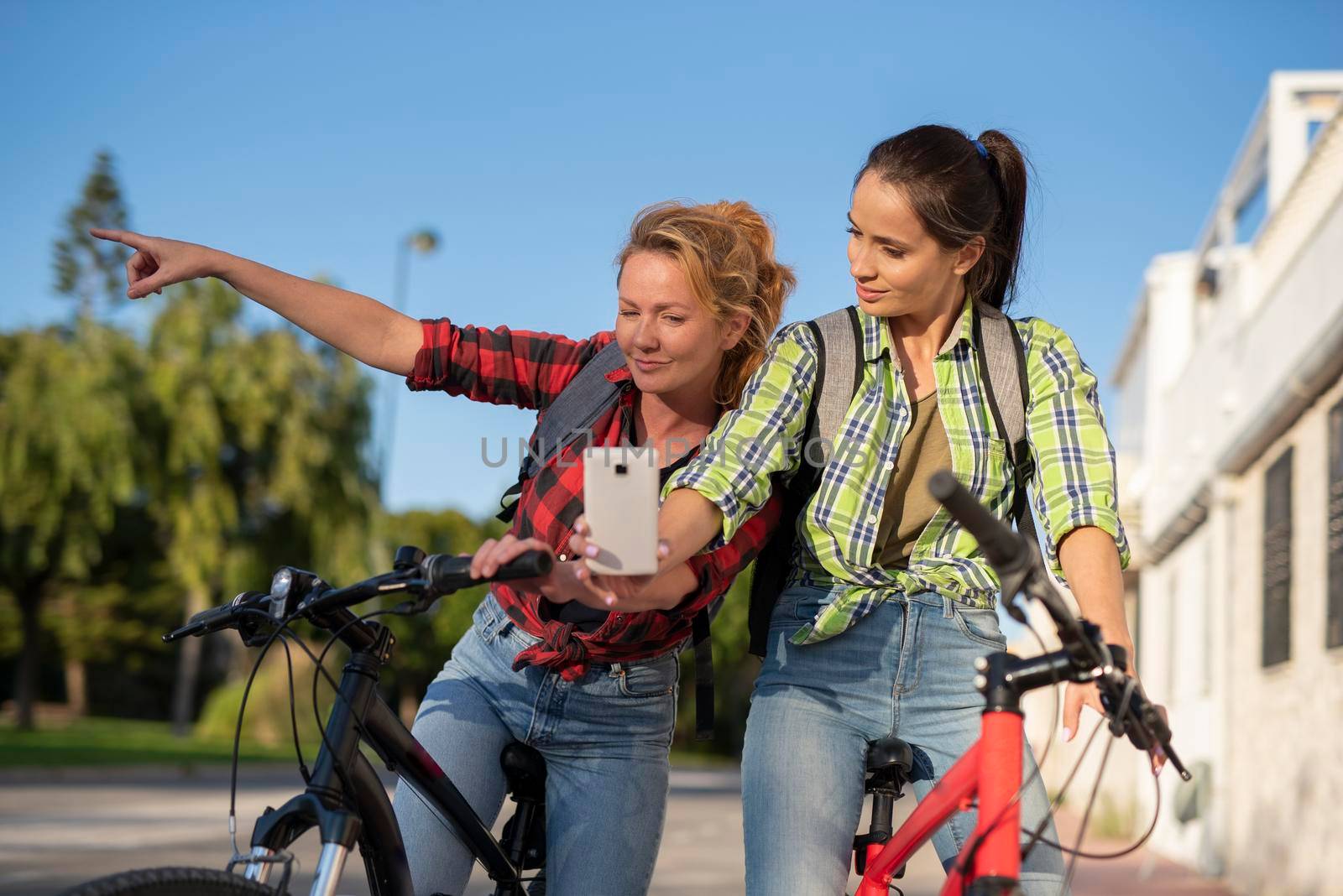 Two pretty young caucasian girls searching the way on a bicycles with smartphone. Best friends enjoying a day on bikes. Sunny summer evening by Alexxoma