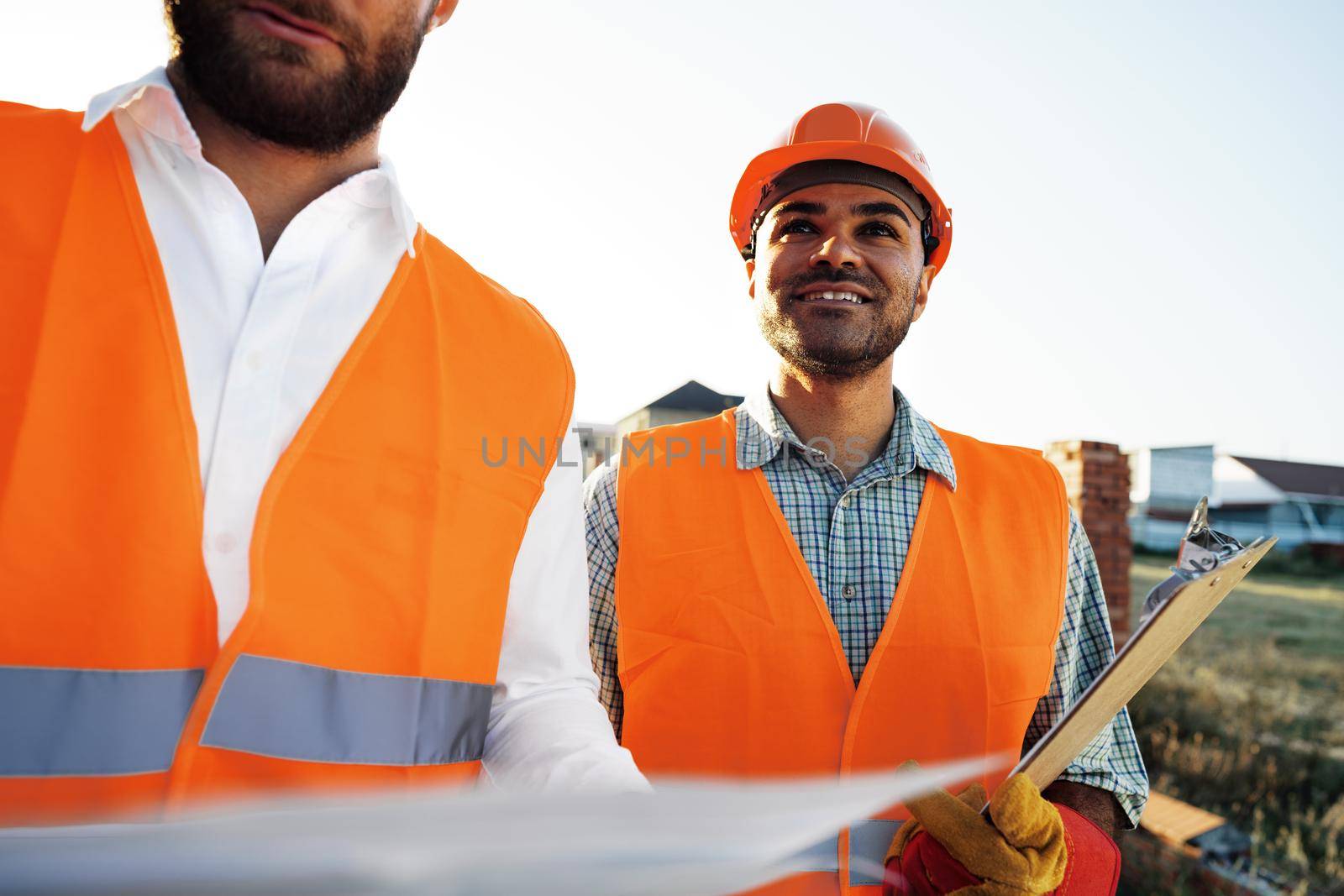 Two young male engineers in uniform and hardhats working at construction site, close up