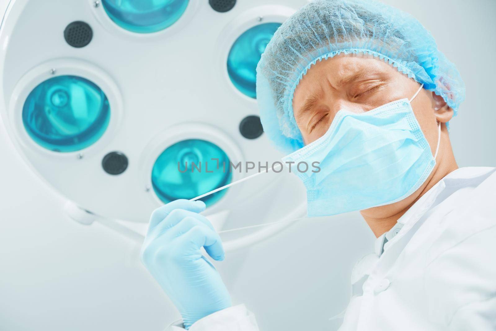 Male surgeon doctor with closed eyes takes off his protective mask after operation