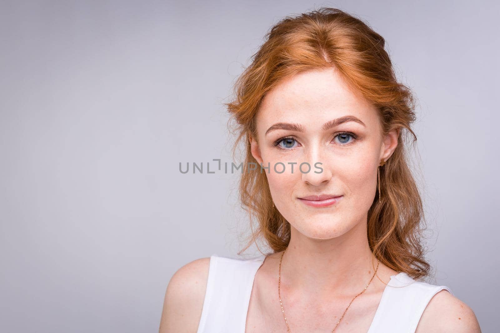 Closeup portrait young, beautiful business woman, student with lred, curly hair and freckles on face on gray background in the studio. Dressed in white blouse with short sleeves about open shoulders.