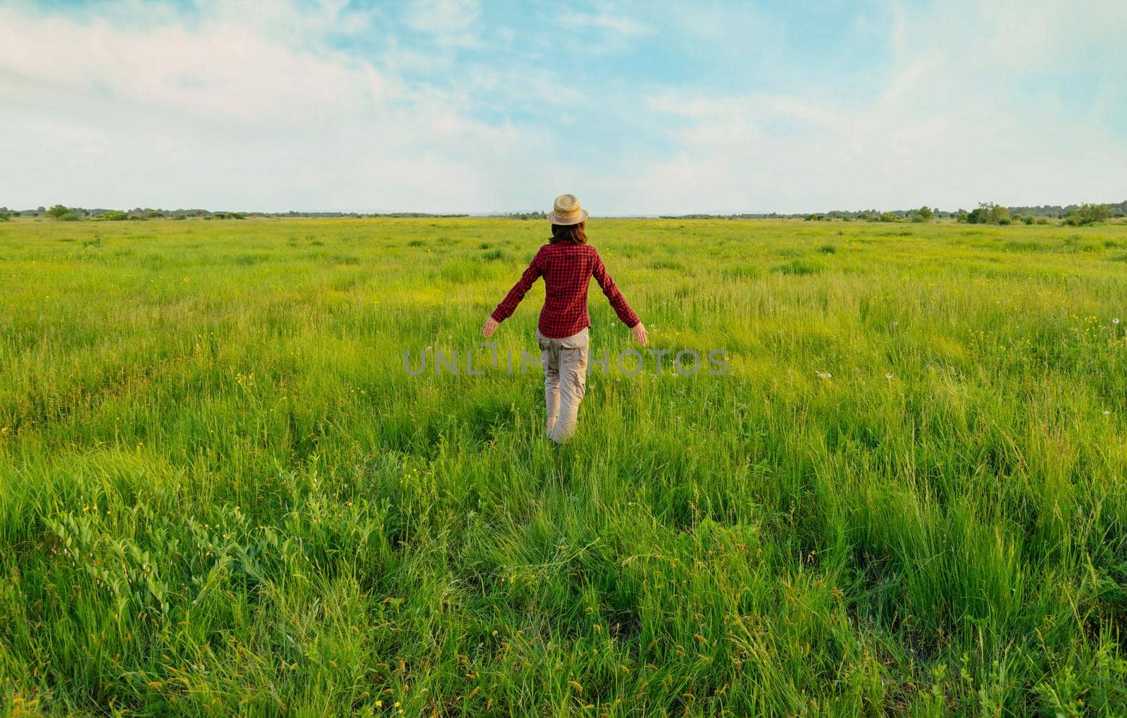 Unrecognizable hipster girl in straw hat walking on summer meadow, rear view. Concept of happiness and carefree