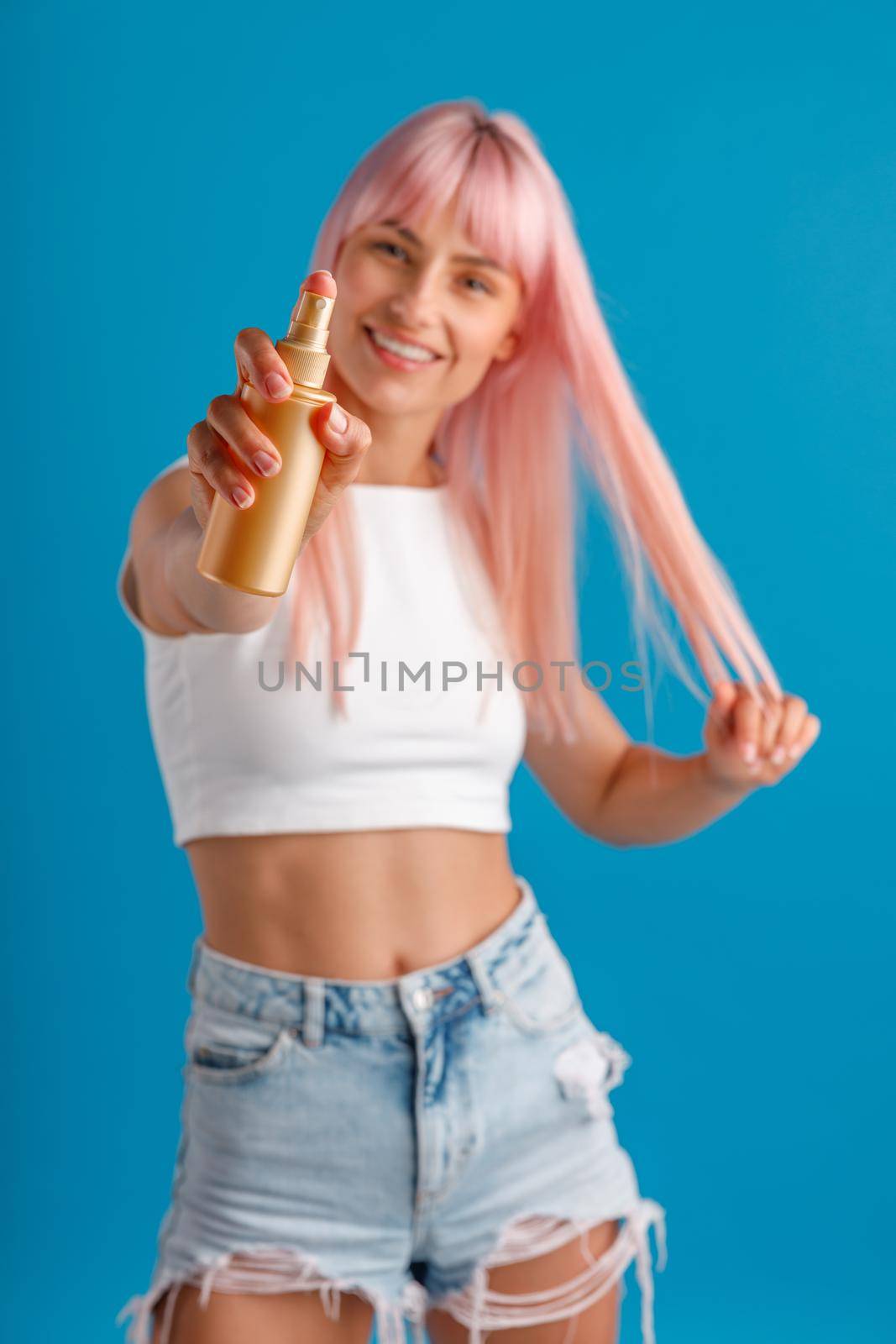 Cheerful young woman with pink hair smiling and showing moisturizing spray to camera while standing isolated over blue studio background by Yaroslav_astakhov