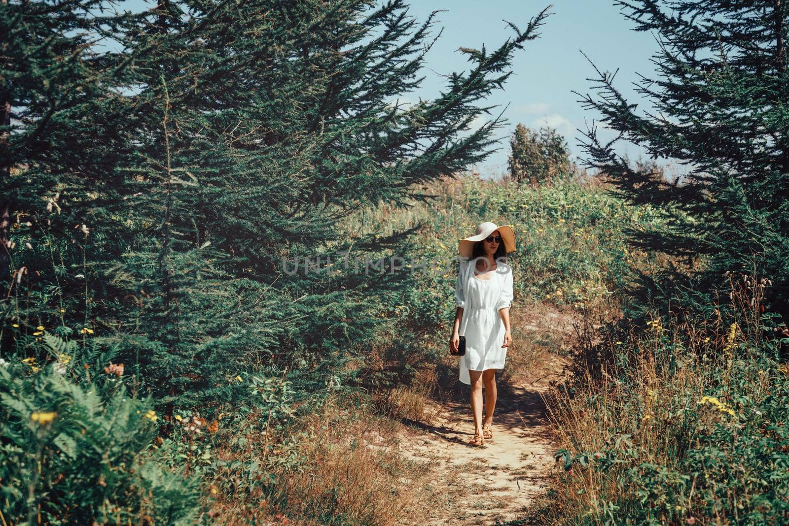 Beautiful young woman in yellow dress and hat with wide brim walking on footpath in summer park