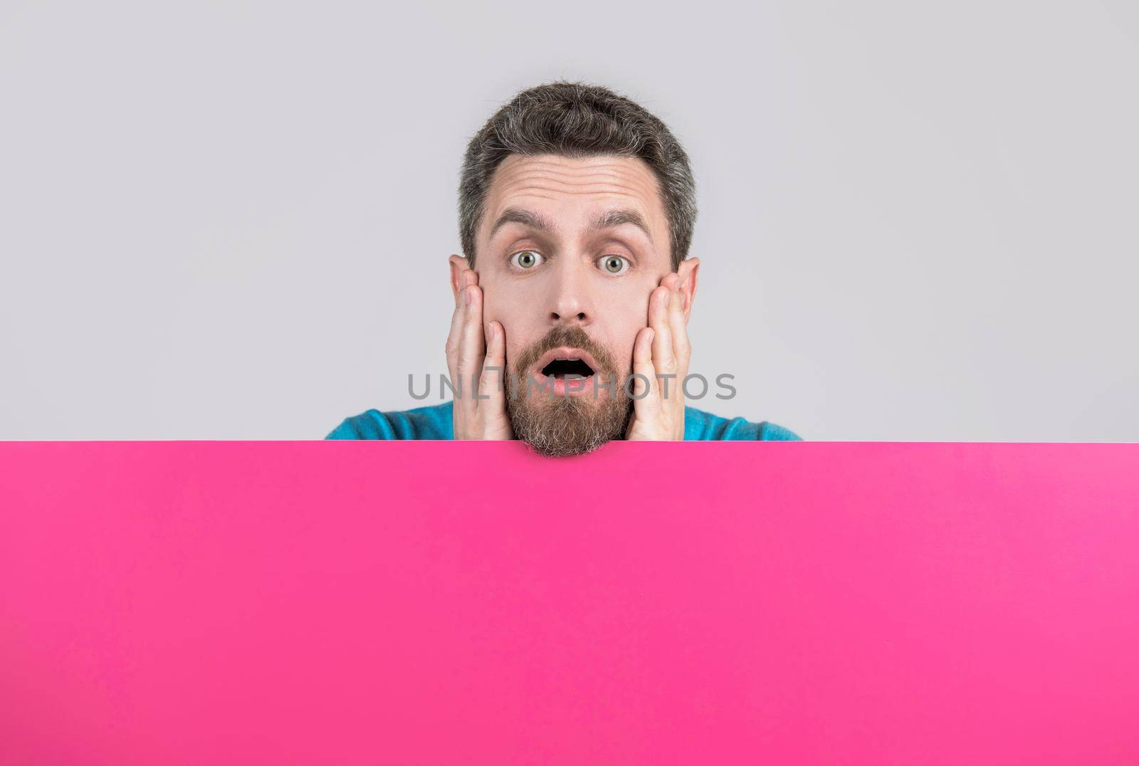 surprised mature bearded man behind blank pink paper banner with copy space for news, discount.
