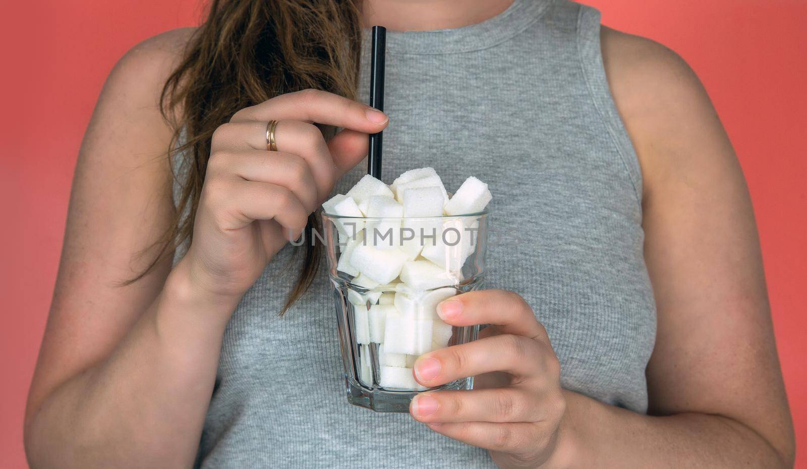 Young woman holding a glass filled with sugar cubes and a straw on pink background, Sweets,diet,fast food,unhealthy eating concept closeup