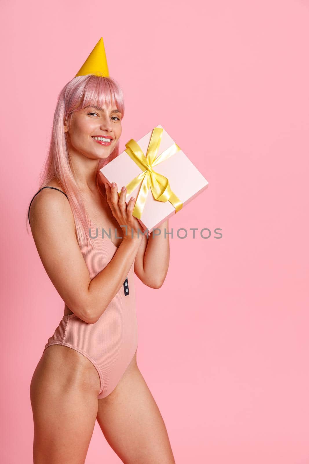 Happy young woman with pink hair wearing swimsuit and party hat holding cute gift box, standing isolated over pink background. Holiday, birthday concept