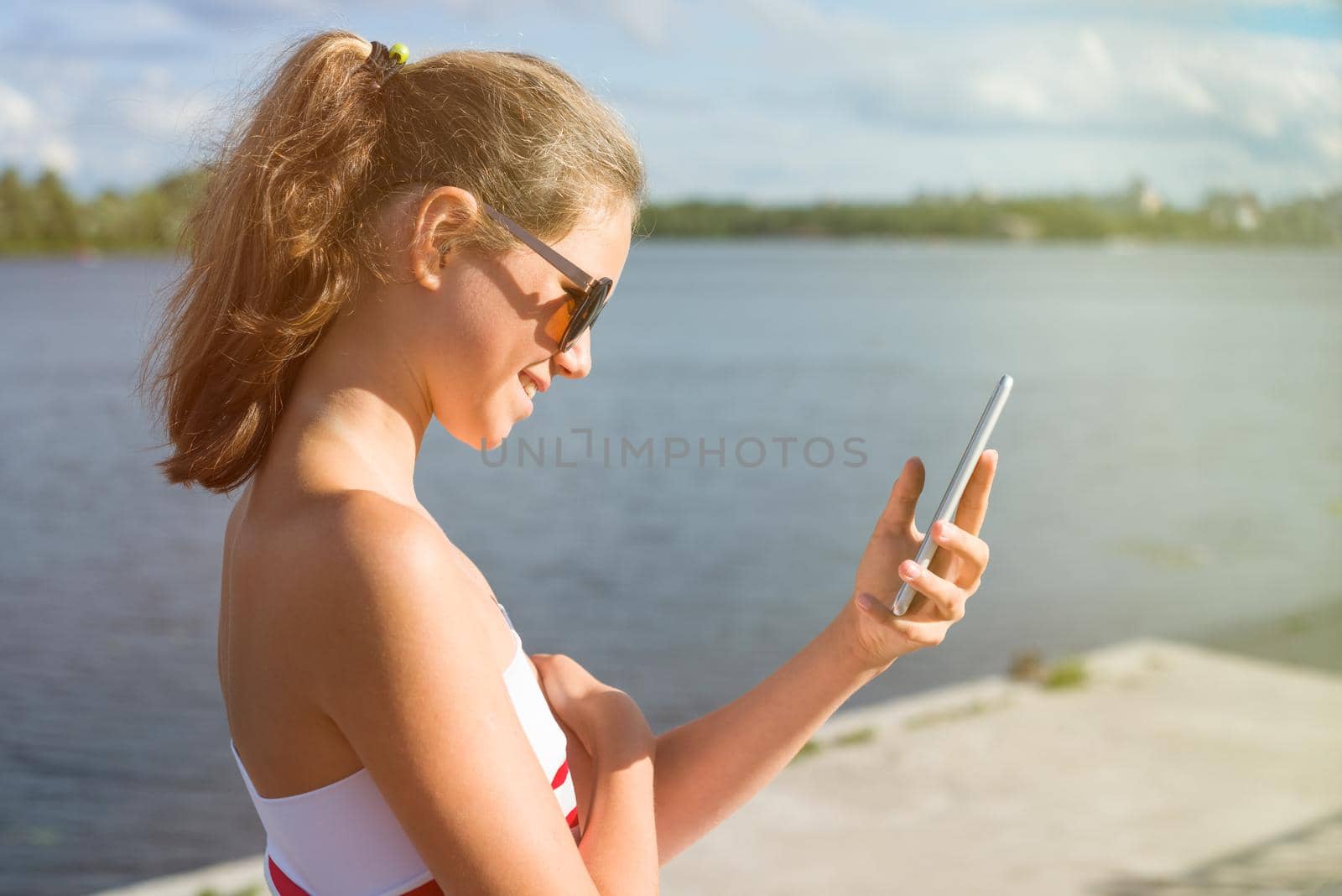 Beautiful young woman in the park using cellphone.In sun glasses, against the background of water, sunset
