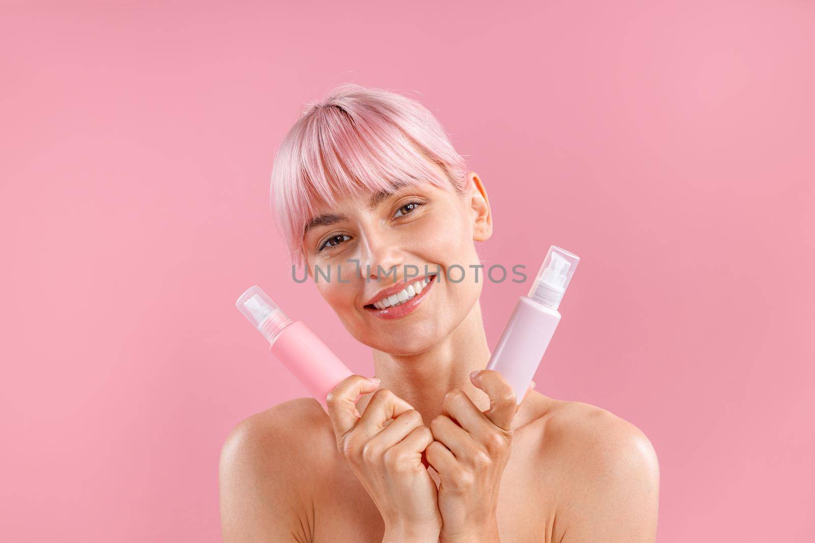 Portrait of lovely young woman with pink hair holding two plastic bottles with beauty products, posing isolated over pink background. Beauty, spa, body care concept