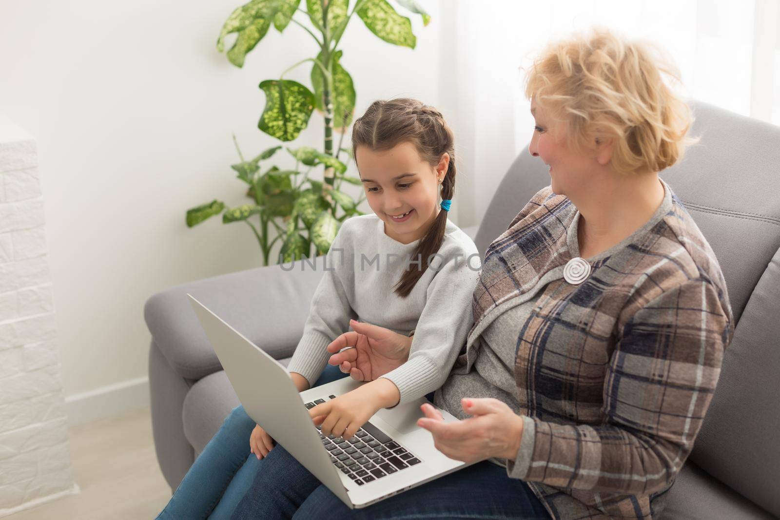 Smart little girl grandchild sit on couch shopping online with excited senior grandmother, teen granddaughter and smiling granny buy on internet browsing laptop relaxing at home together