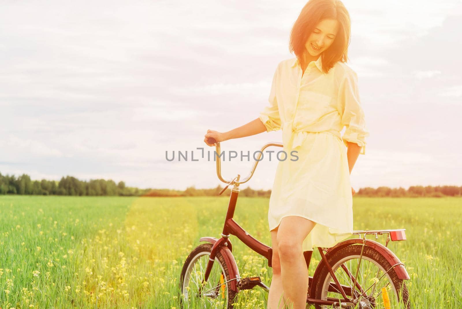 Beautiful young woman in dress walking with bicycle on summer meadow at sunny day. Image with sunlight effect