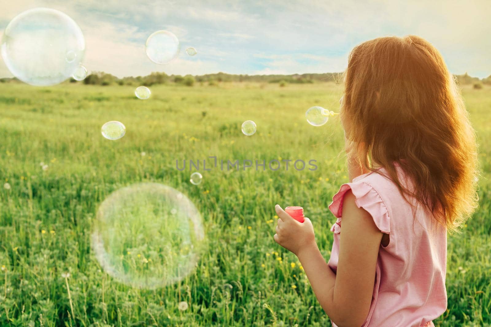 Little girl in pink dress blowing soap bubbles on summer meadow at sunny day, rear view. Image with sunlight effect