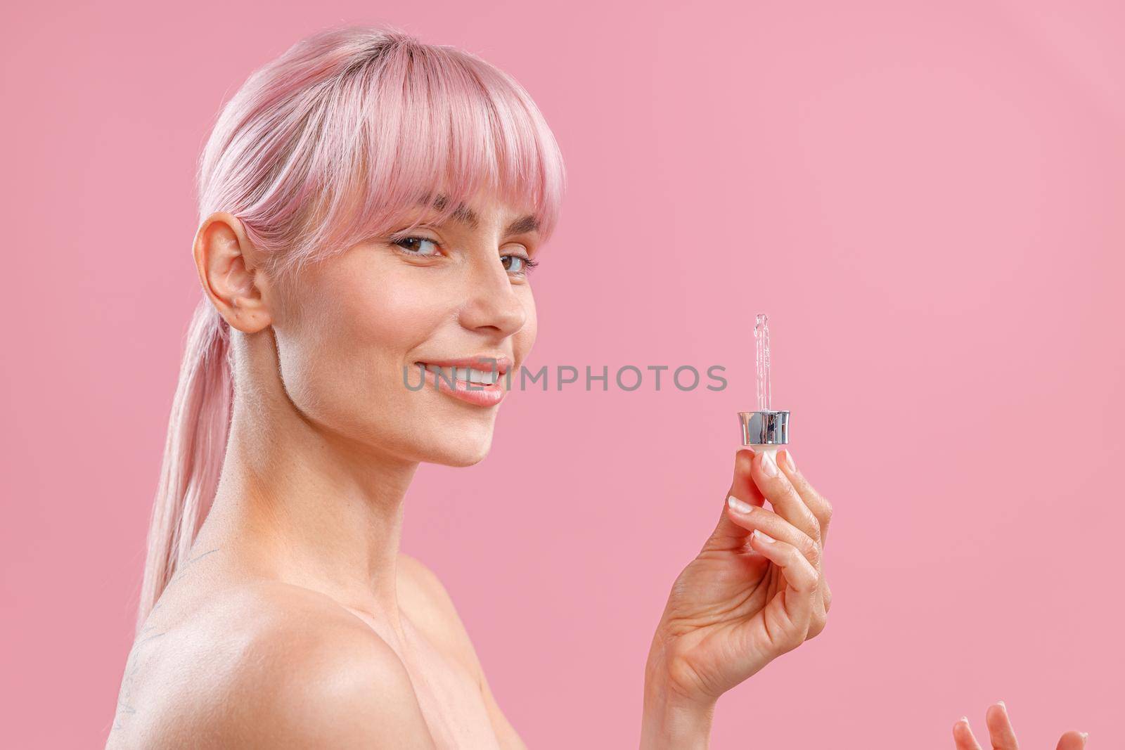 Beautiful young woman with pink hair and perfect skin smiling at camera, holding dropper with serum or hyaluronic acid, posing isolated over pink background. Beauty, cosmetics and skincare. Side view