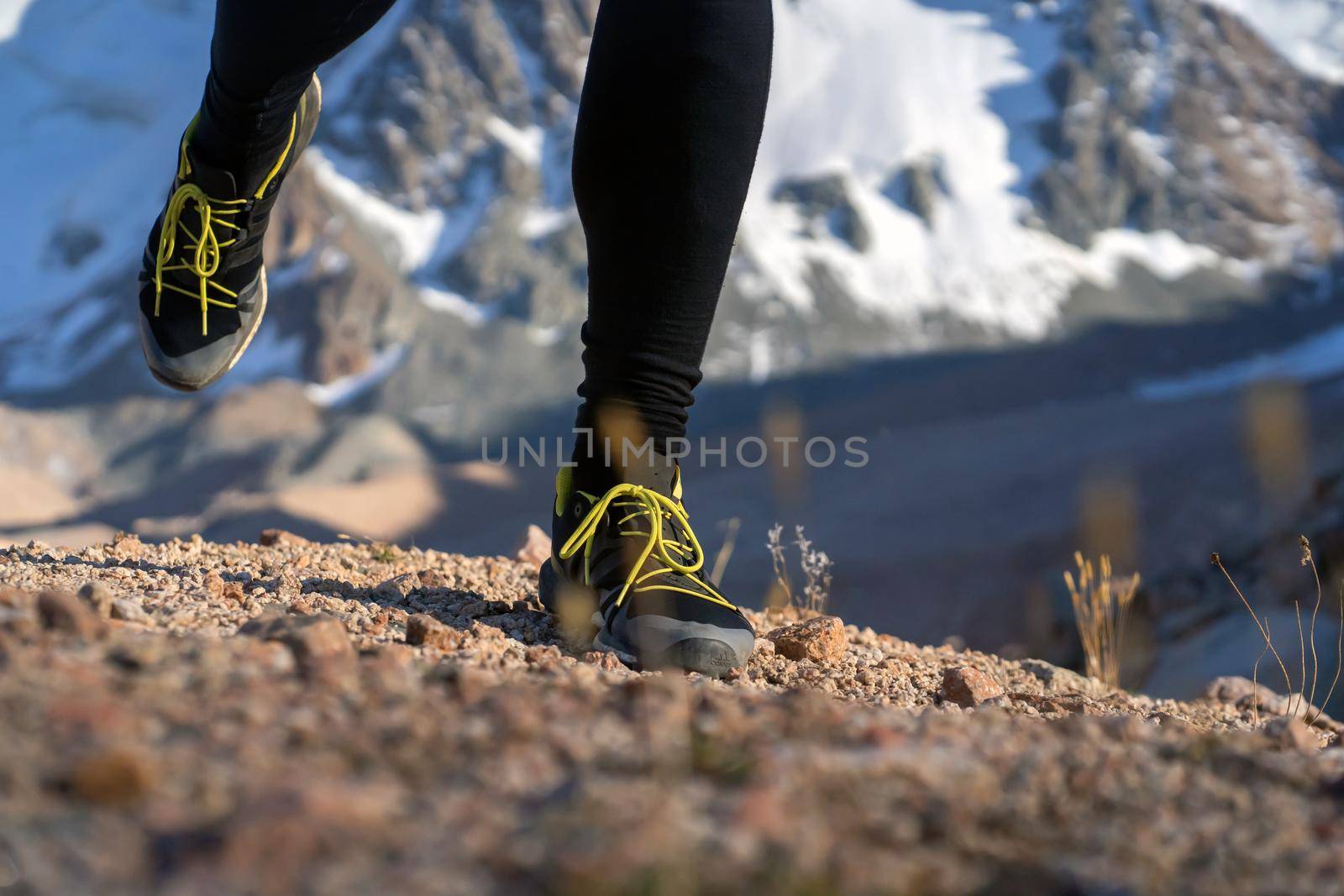 Athlete runner legs run on a trail along a rocky path close-up. Fitness and warm-up, young man with snow-covered mountain peaks in the background and open space around him at sunset.