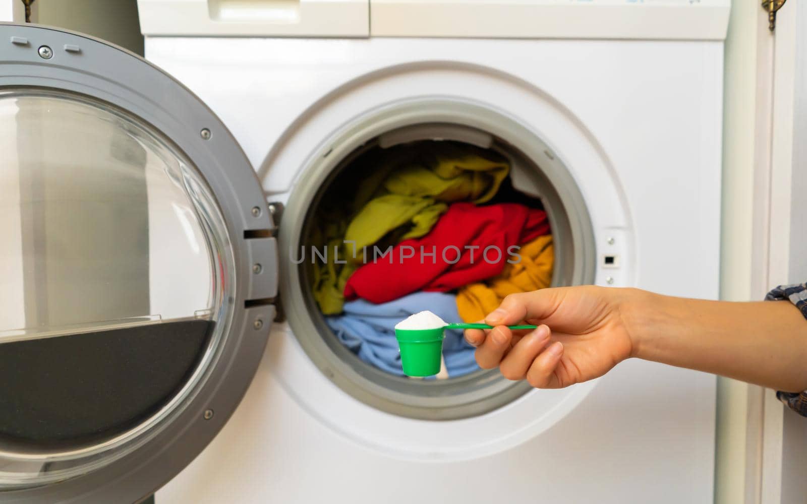 A woman's hand holds a cap with washing powder against the background of a washing machine with bright things put into it. The girl washes and wringes out things in the laundry room at home.