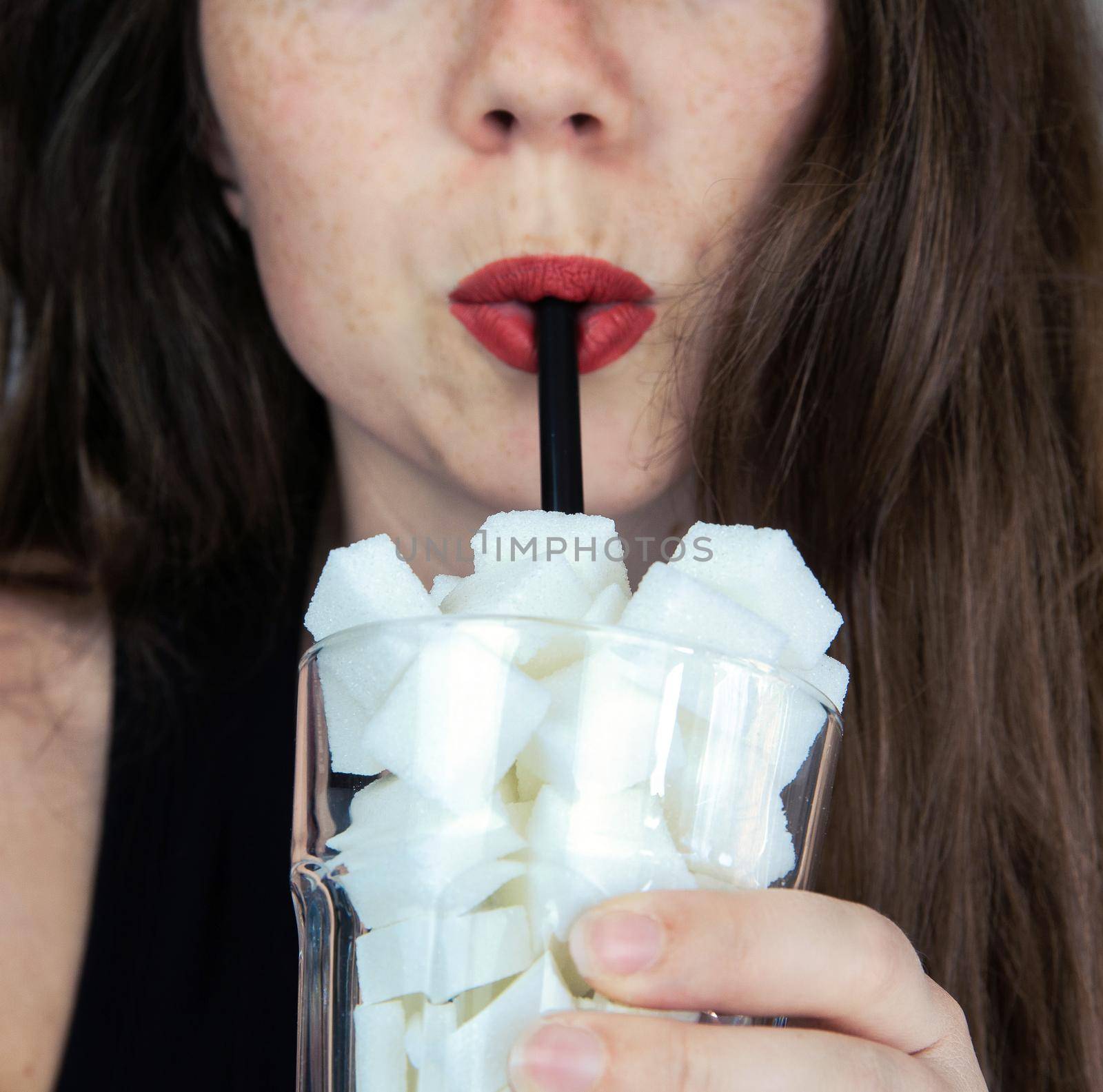 Portrait of young woman drinking with a black colored straw from a glass filled with sugar cubes Junk food, unhealthy diet, too much sugar on drinks, nutrition concept closeup