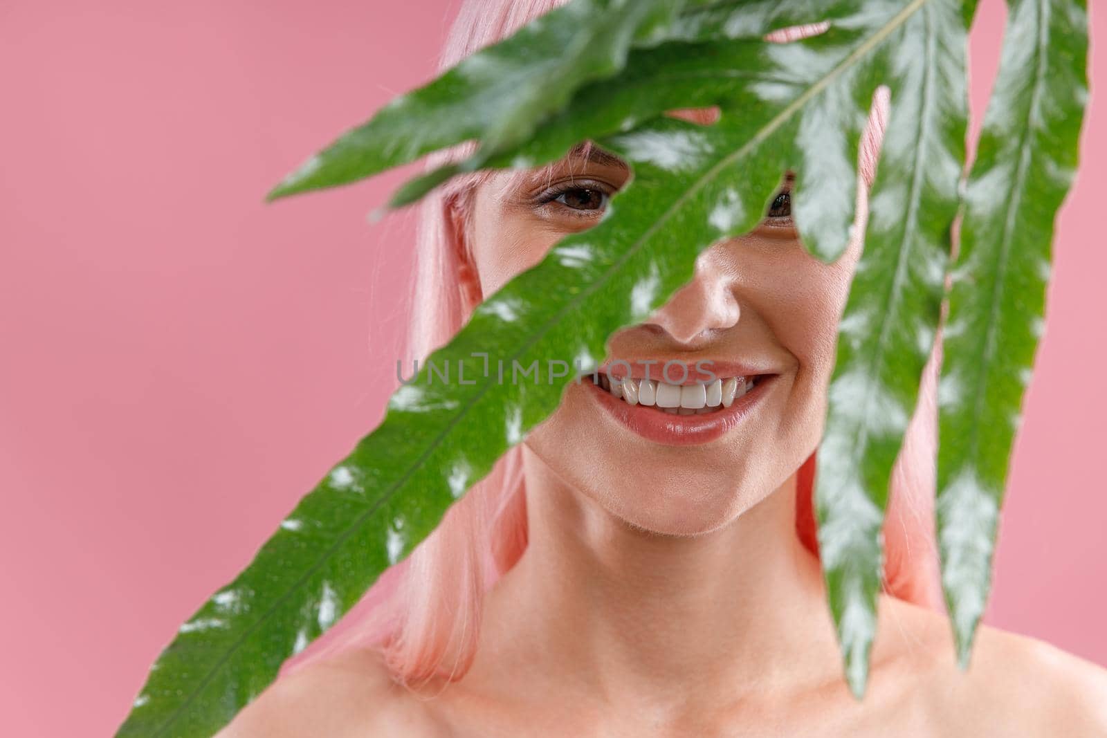 Portrait of happy woman with pink hair smiling at camera, hiding behind green plant leaf, posing isolated over pink studio background. Beauty, skin care, natural cosmetics concept