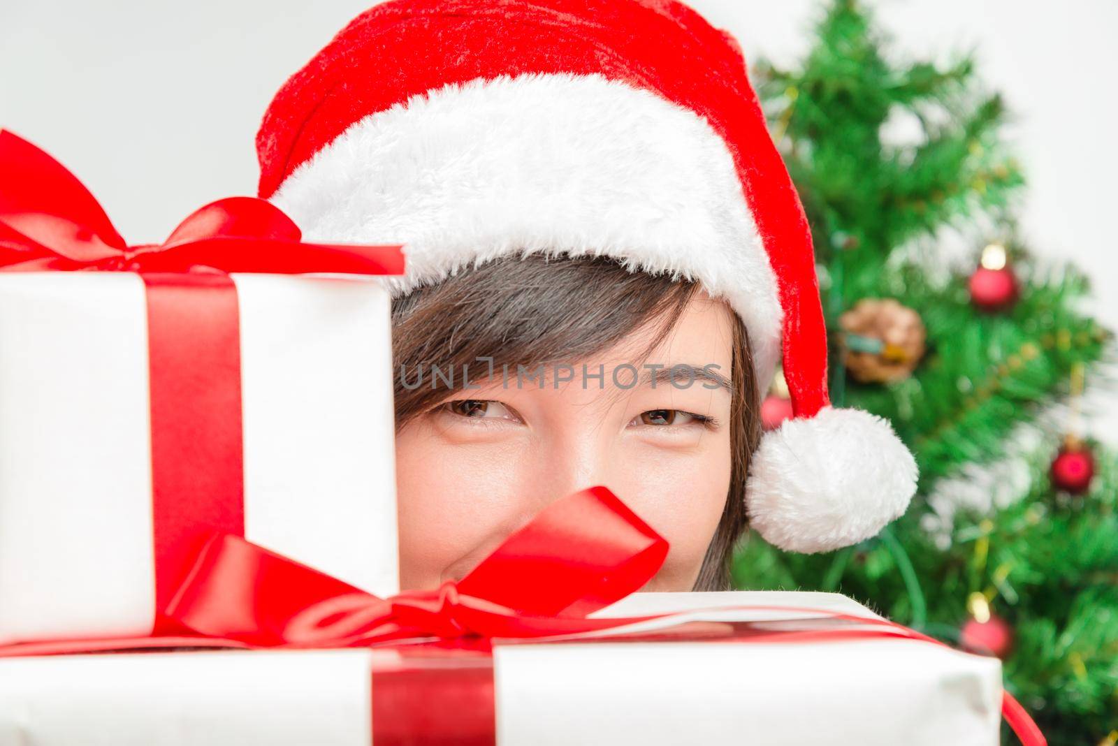 Young woman in santa hat looks out from behind presents on a background of Christmas tree