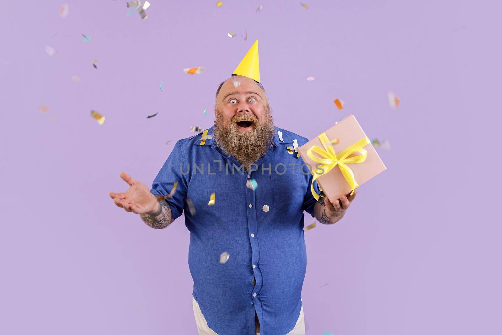 Positive emotional plus size man in tight blue shirt and party hat holds gift box standing under confetti shower on purple background in studio