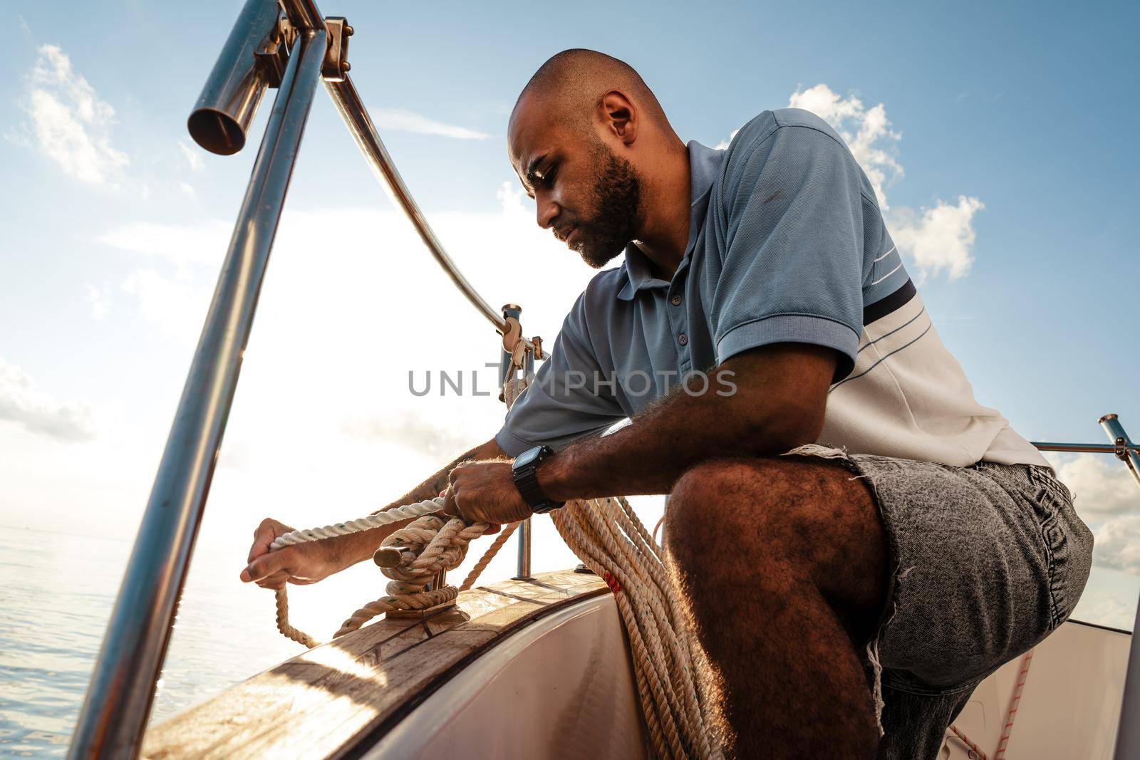 Young african american sailor tying ropes on sailboat in the sea on sunset, close up