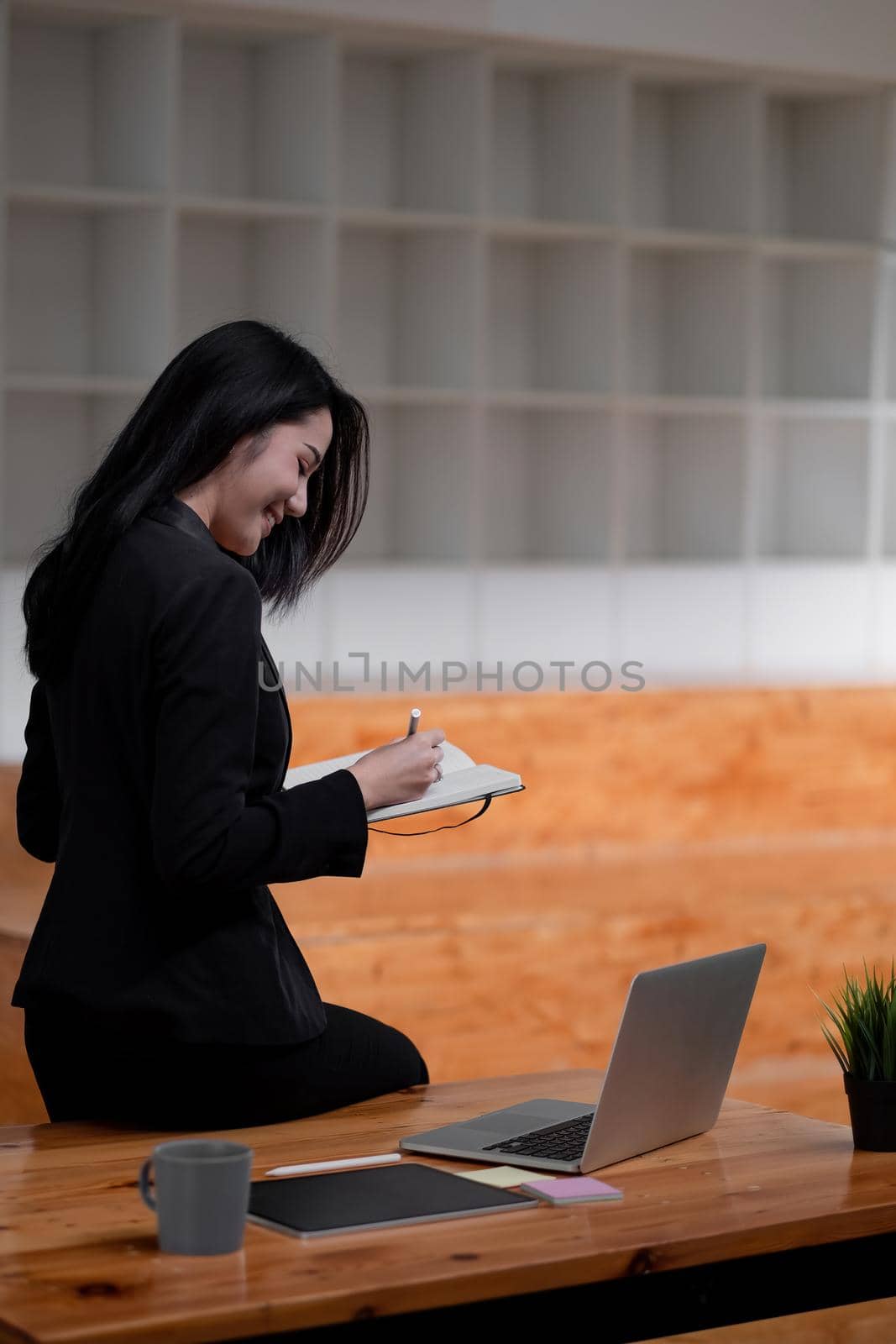 Asian business woman working at office and taking note, Female sitting on desk and using laptop, tablet.