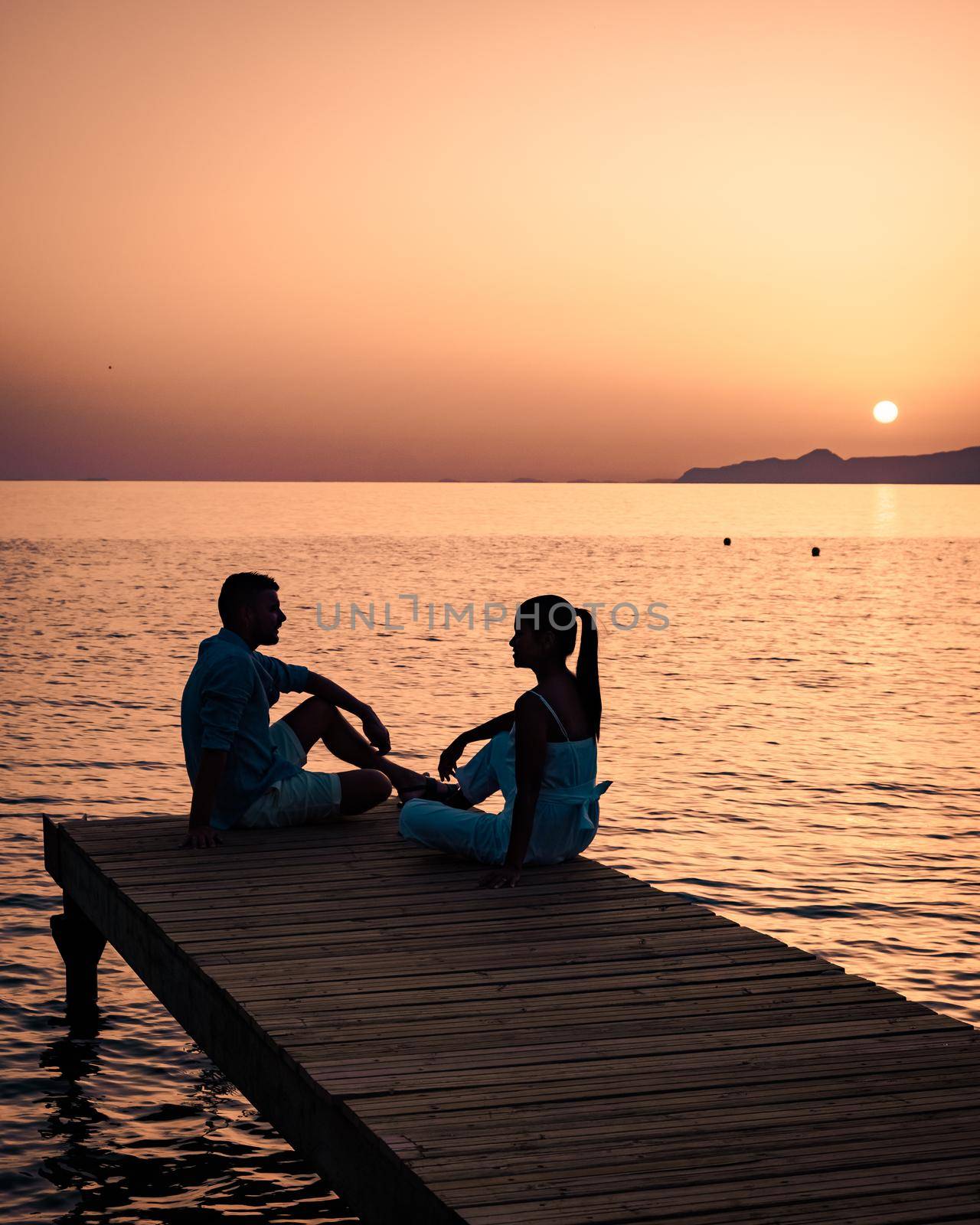 a couple seated on a wooden jetty, looking at colorful sunset on the sea, men, and women watching a sunset in Crete Greece Europe