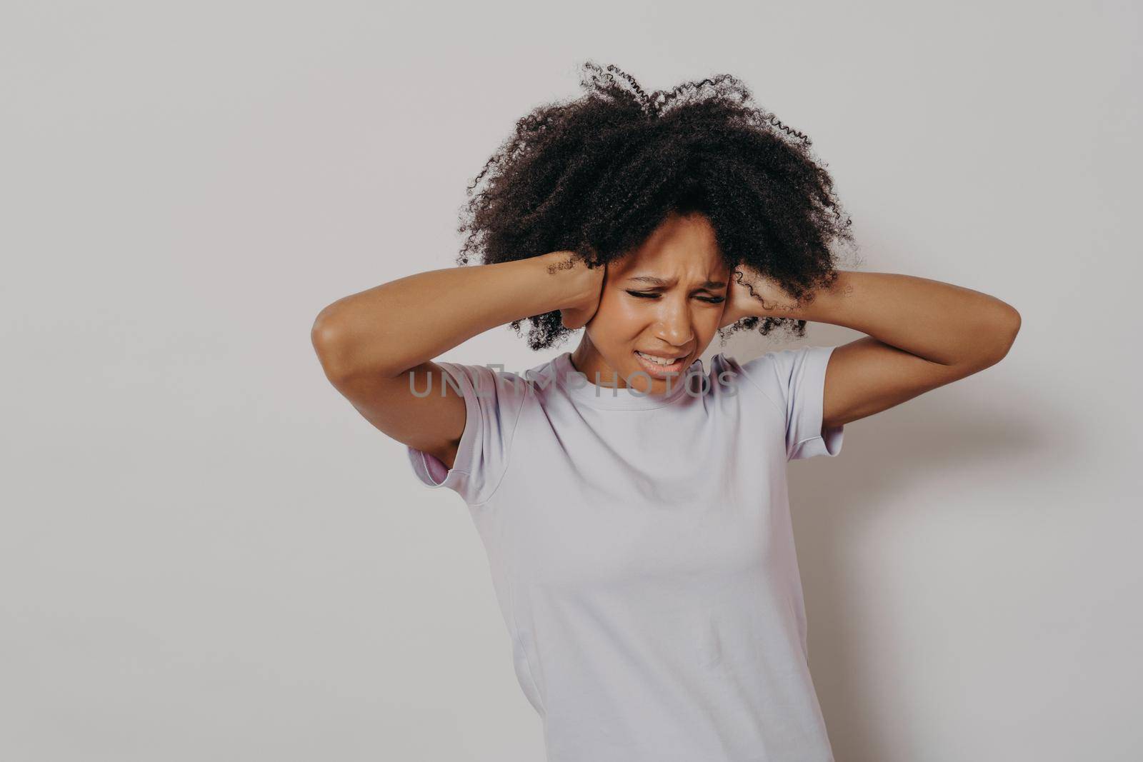 Studio portrait of young angry annoyed african woman covering her ears with both hands to avoid loud noise while posing against white background, dressed in white tshirt. Negative human emotions