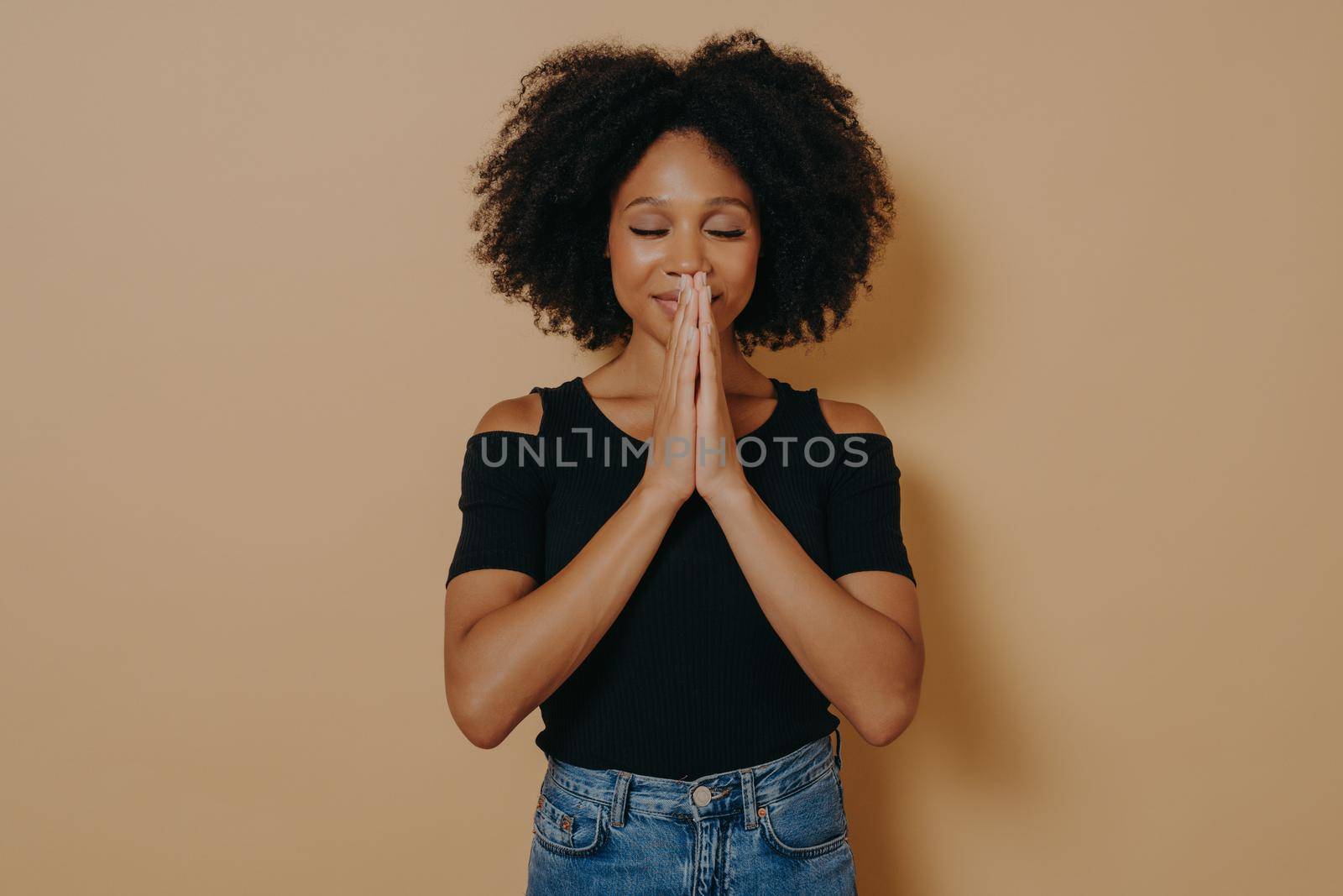 Afro American young woman praying and holding hands in prayer gesture standing in studio by vkstock