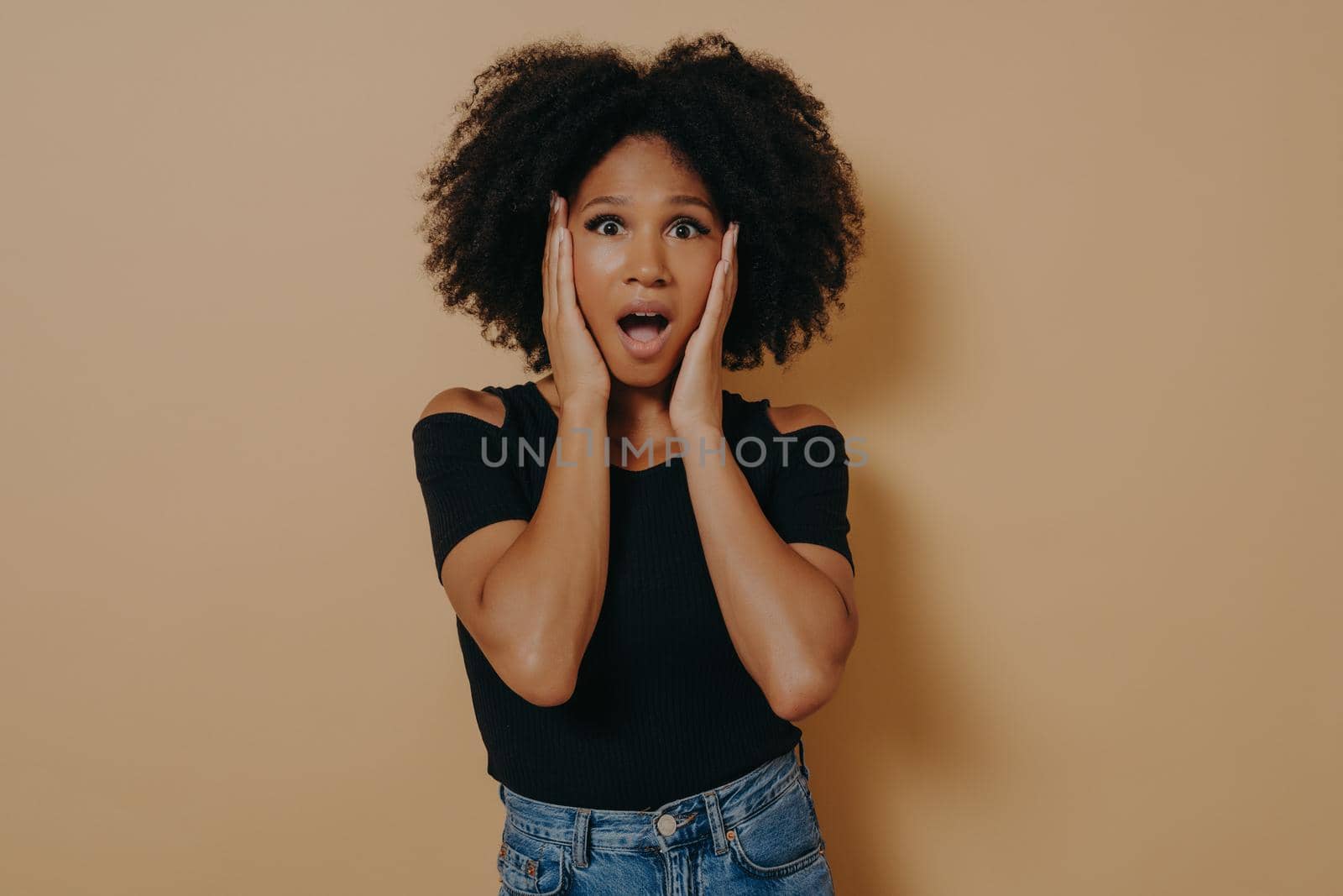 Photo of shocked pretty dark skinned curly haired lady holding hands on cheekbones by vkstock