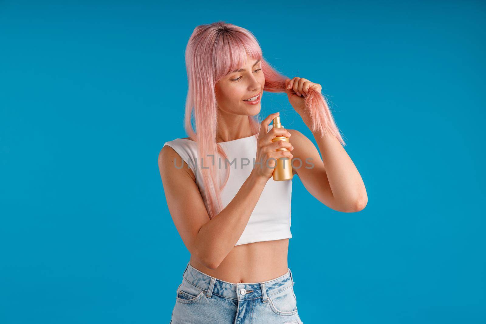 Caring young woman smiling and applying moisturizing spray to her smooth pink hair while standing isolated over blue studio background. Beauty products, hair care concept