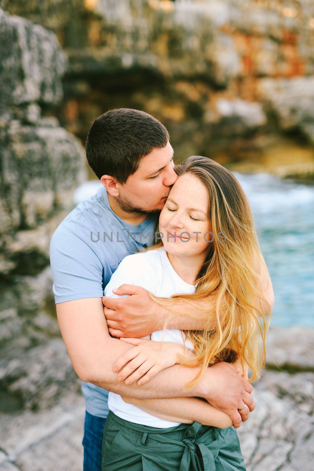 Husband hugs from behind and kisses smiling wife against the background of a rocky shore and water. High quality photo