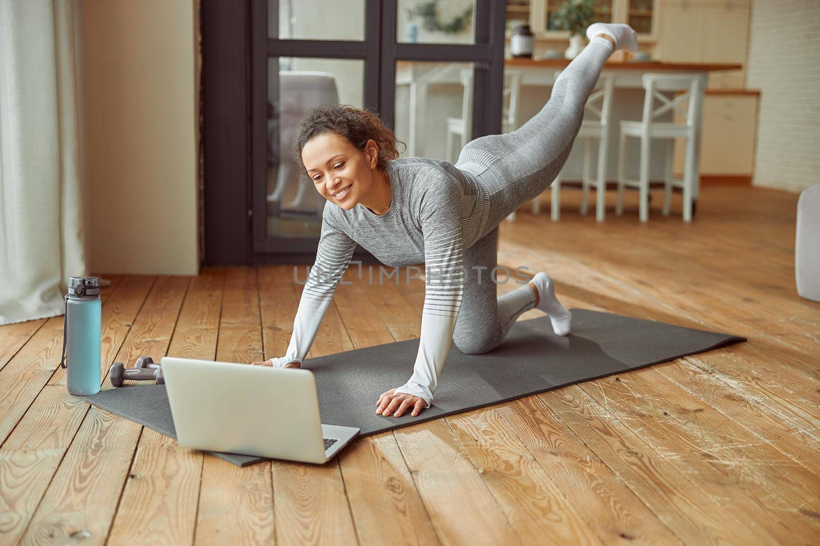 Happy slim female is exercising on mat while watching workout video on notebook in living room